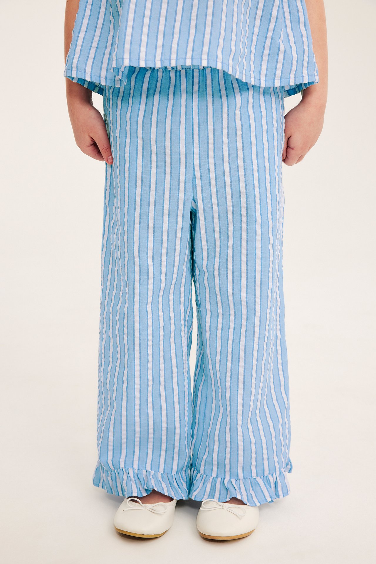 Framifrån: barn i blåvitrandiga bäckeböljebyxor med volanger, matchande topp och vita ballerinaskor.