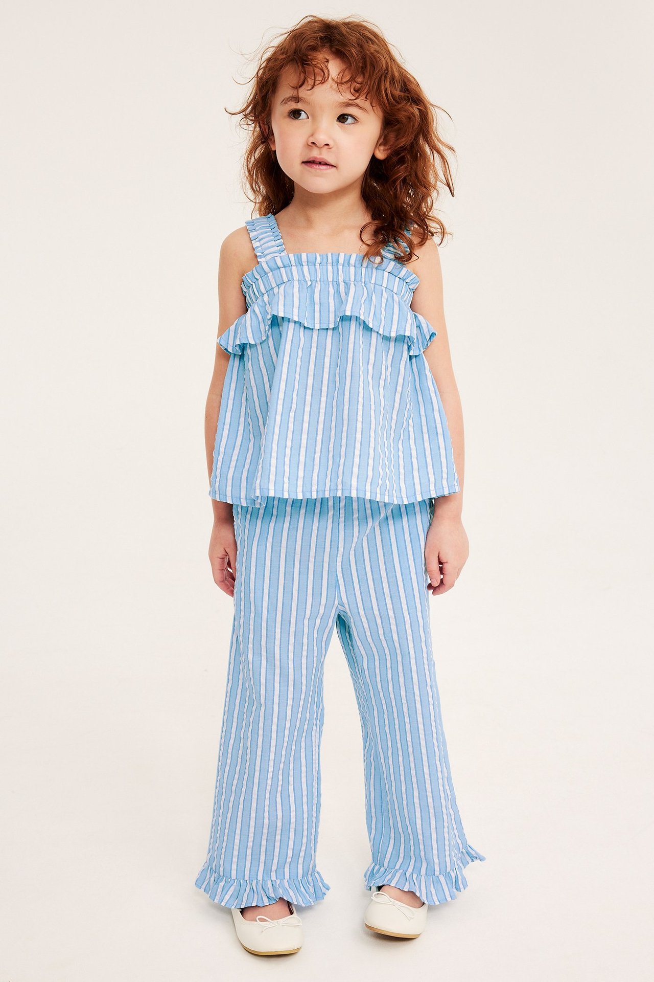 Lapsi edestä: siniset raidalliset seersucker-housut röyhelöillä, yhteensopiva toppi ja valkoiset kengät.