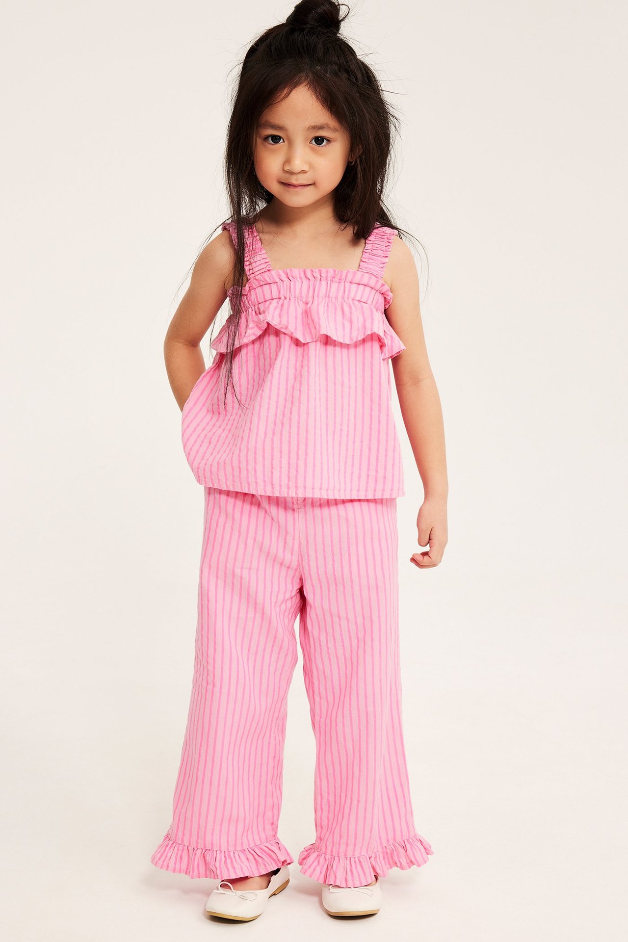 Framifrån: Kid bär ett matchande rosa randigt bäckebölja-set med volangbyxor och volanglinne.