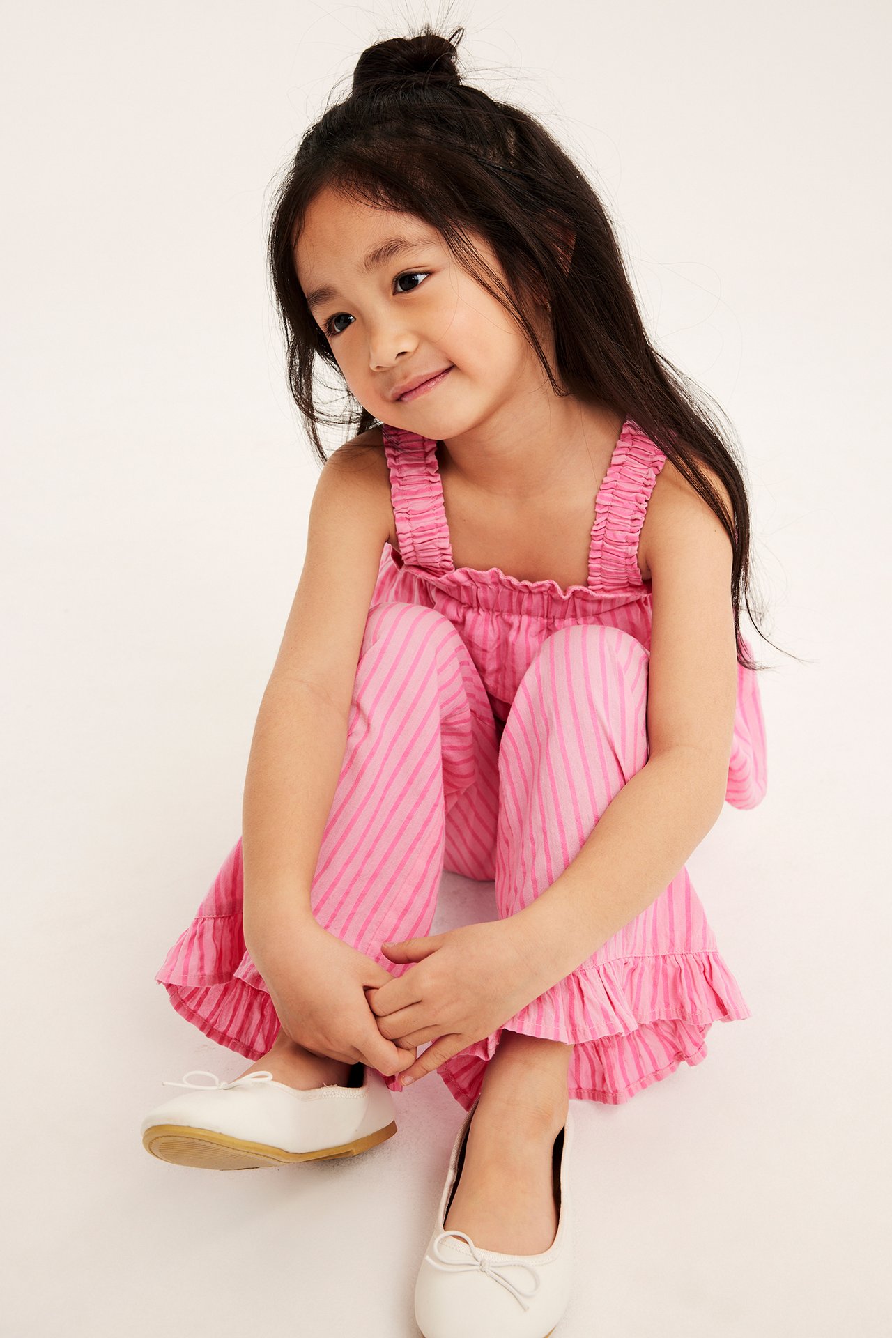 Ett barn bär ljusrosa randiga bomullsbyxor med volangkanter, en matchande topp och vita ballerinaskor.