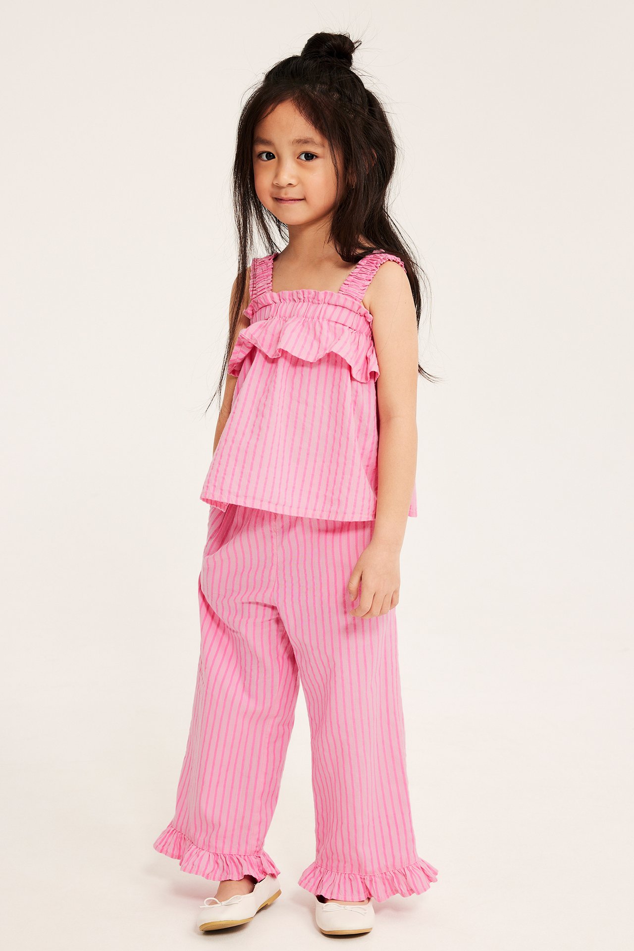 Framsida av ett barn i rosa randig bomullstopp med volanger, matchande byxor och vita skor.