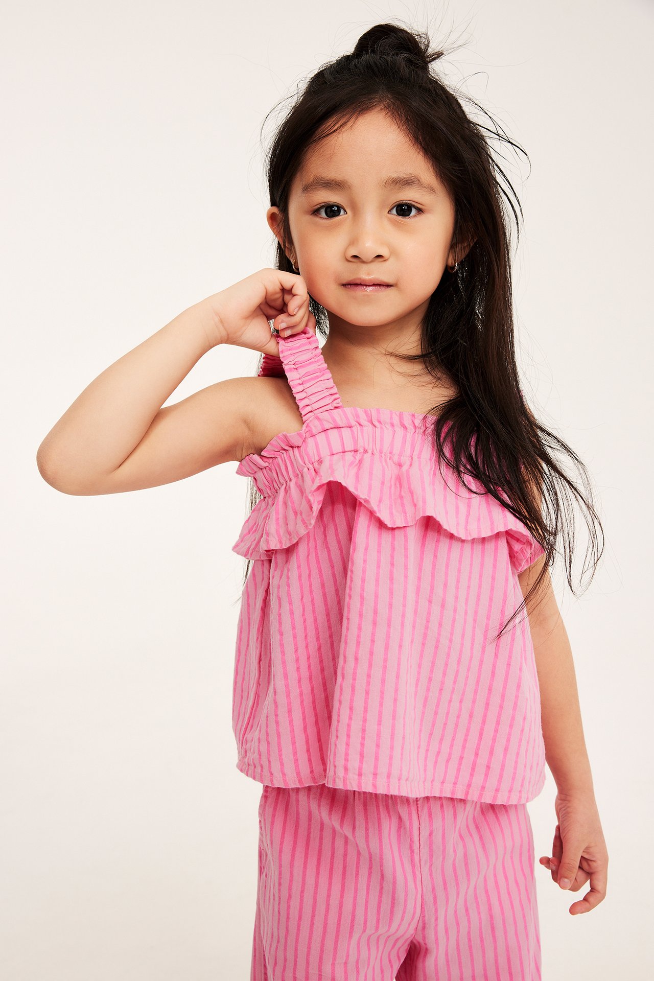 Widok z przodu, dziecko w jasnoróżowej bawełnianej bluzce w paski z falbankami i pasujących spodniach.
