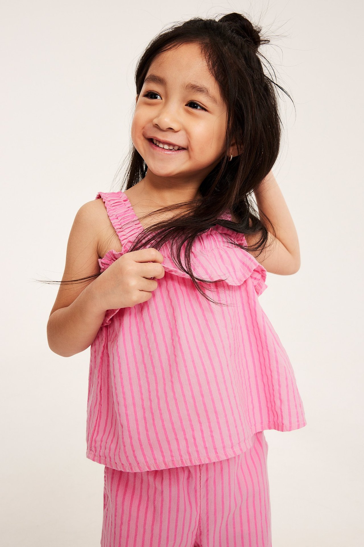 Hymyilevä lapsi edestäpäin yllään vaaleanpunainen raidallinen röyhelötoppi ja yhteensopivat shortsit.