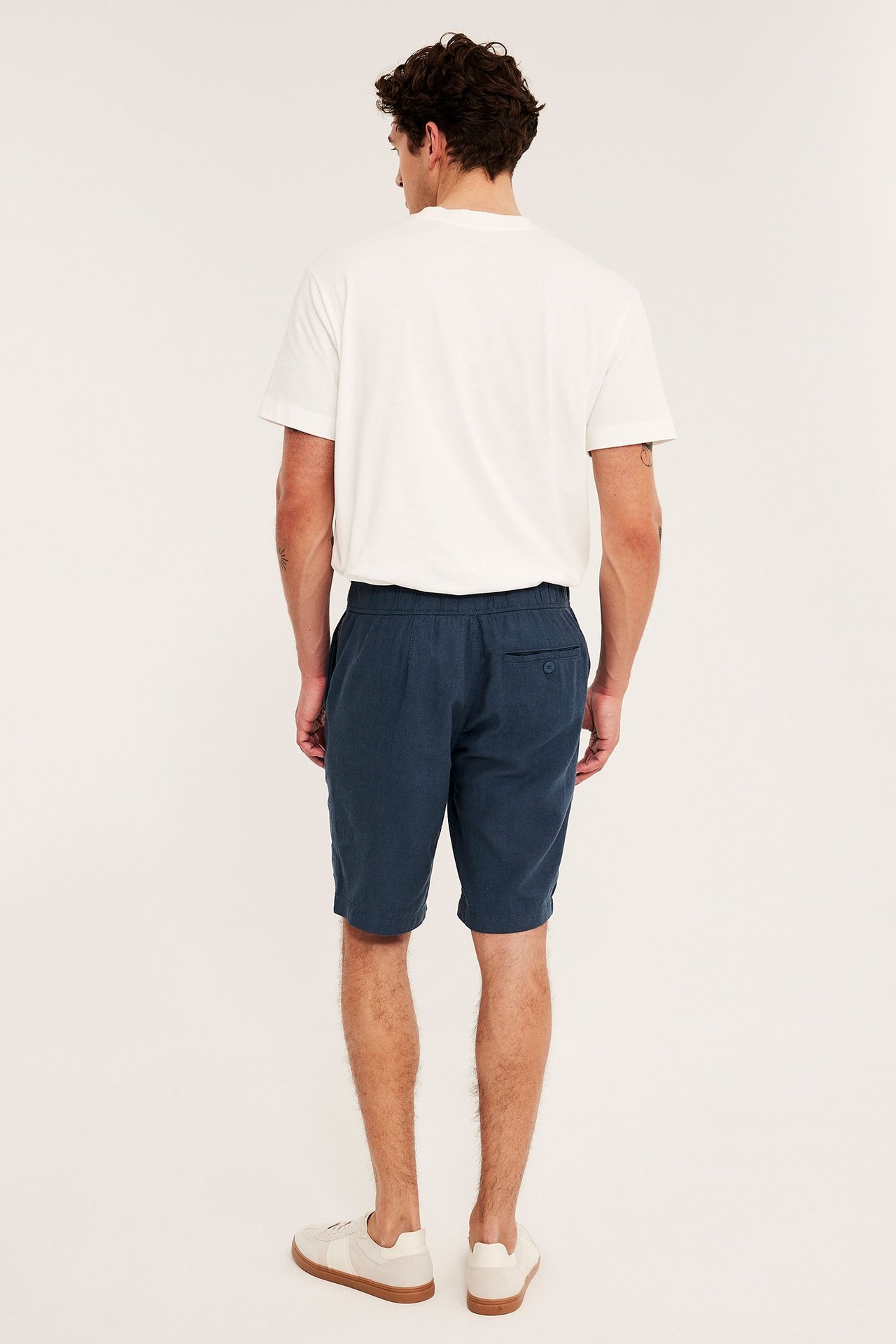 Mężczyzna tyłem: ciemnoniebieskie szorty z mieszanki lnu z elastyczną talią, biały t-shirt i beżowe trampki.