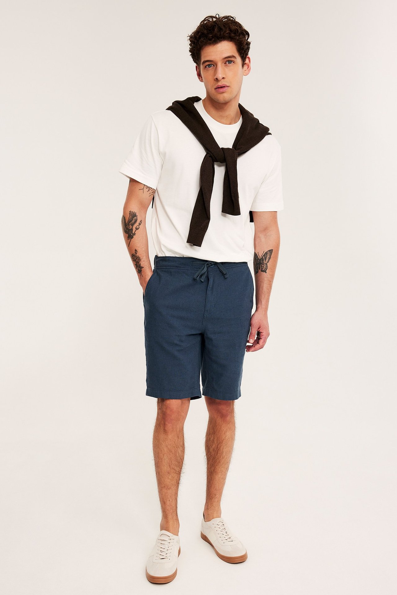 Framifrån, man i mörkblå linneshorts med dragsko, vit t-shirt, mörkbrun tröja över axlarna och beiga sneakers.