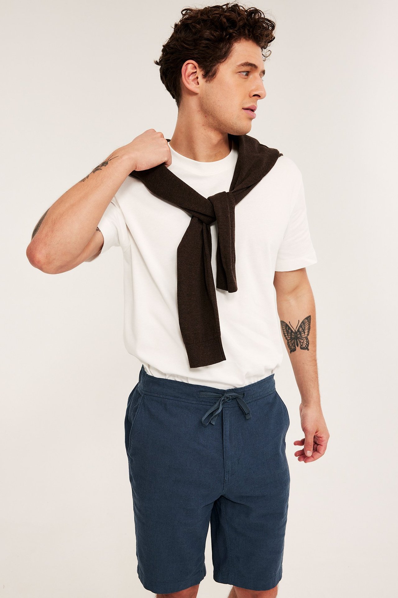 Man framifrån i mörkblå linneshorts med dragsko, vit t-shirt och en brun tröja draperad över axlarna.