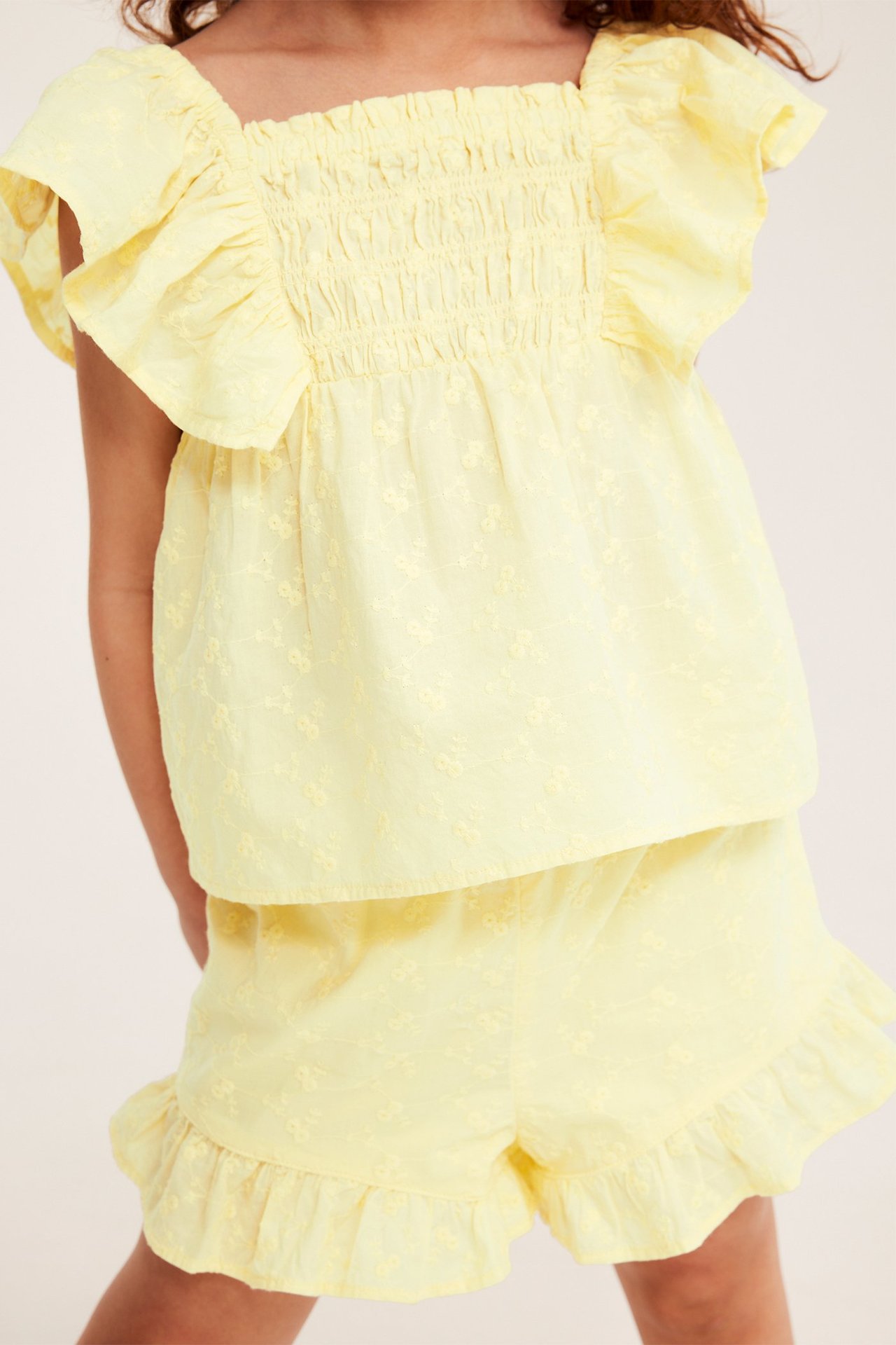 Etunäkymä lapsesta vaaleankeltaisissa kirjailluissa röyhelöshortseissa ja yhteensopivassa smockatussa puuvillatopissa.