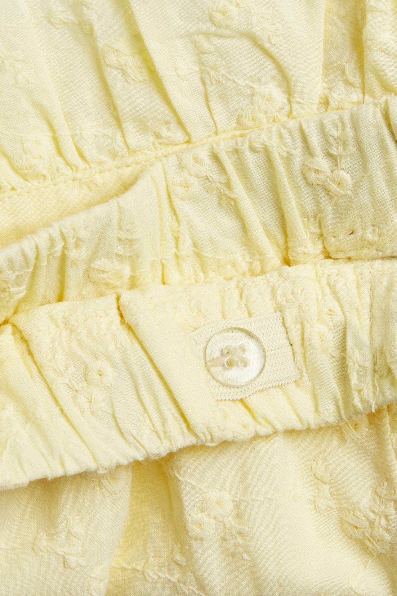 Närbild av ljusgula broderade bomullsshorts med volang och justerbar midja med knapp.