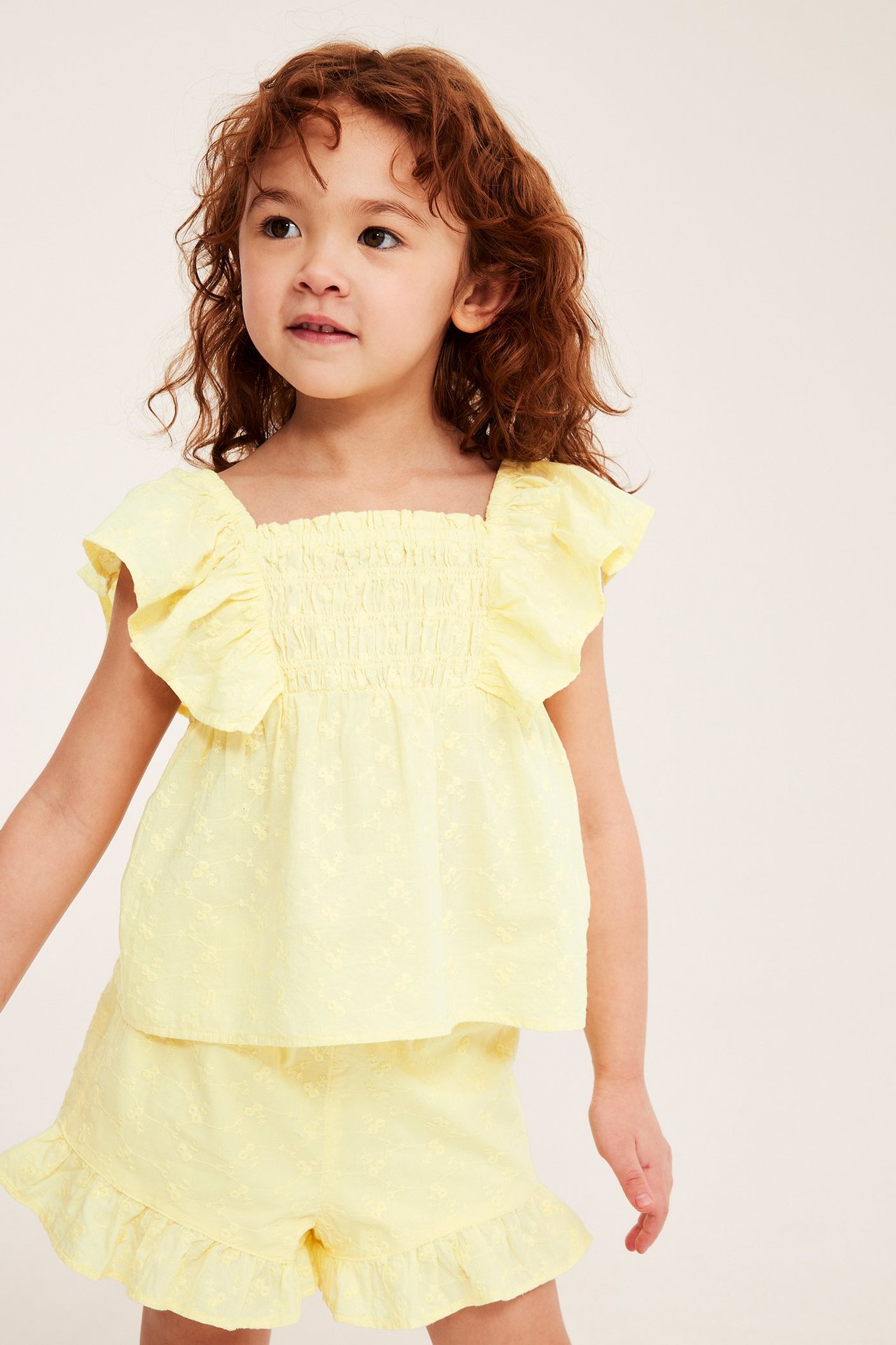 Widok z przodu: dziecko w jasnożółtych haftowanych szortach z falbankami i pasującym topie.