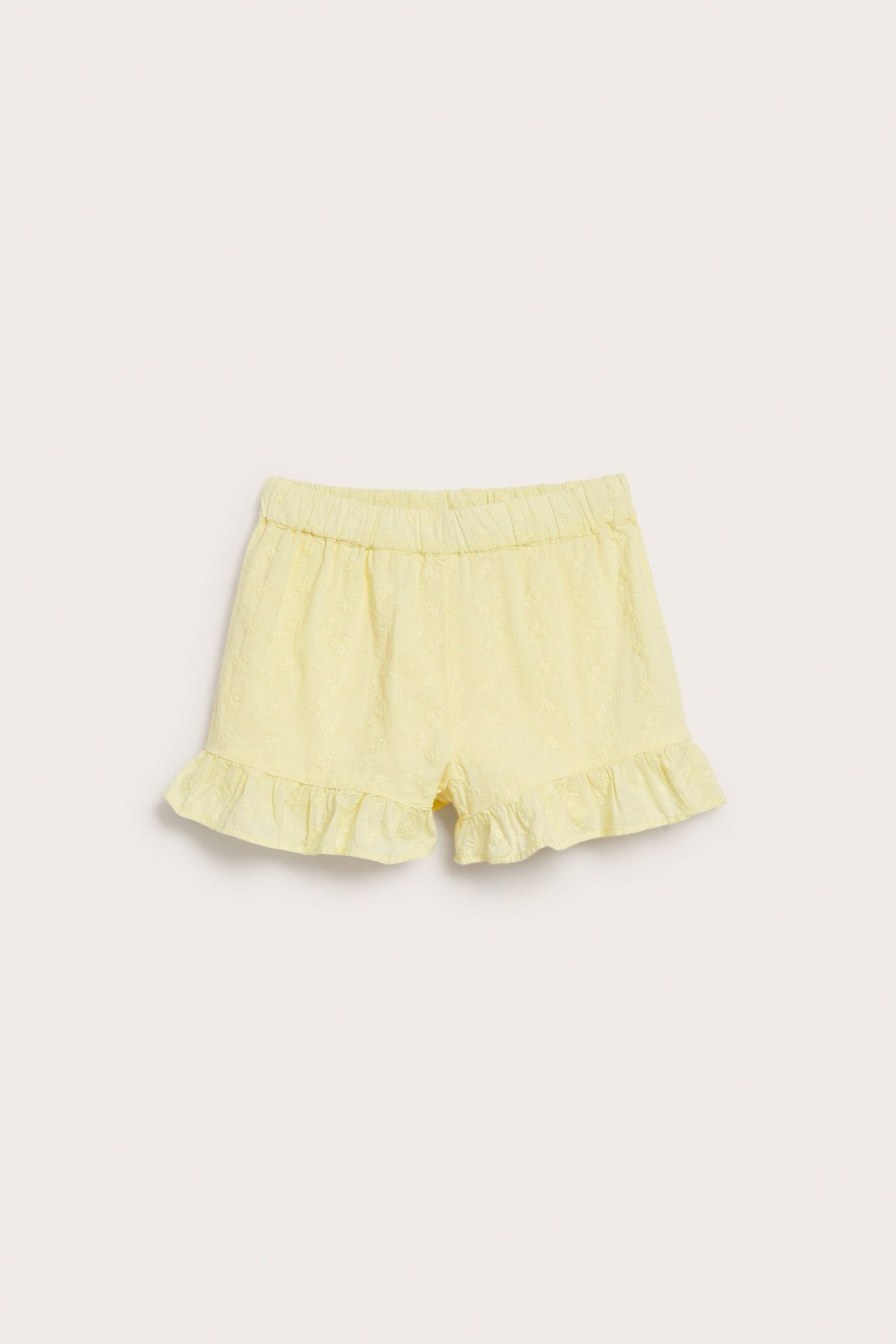 Framsida av ljusgula barnshorts i bomull med diskret brodyr, resårmidja och volangkanter.