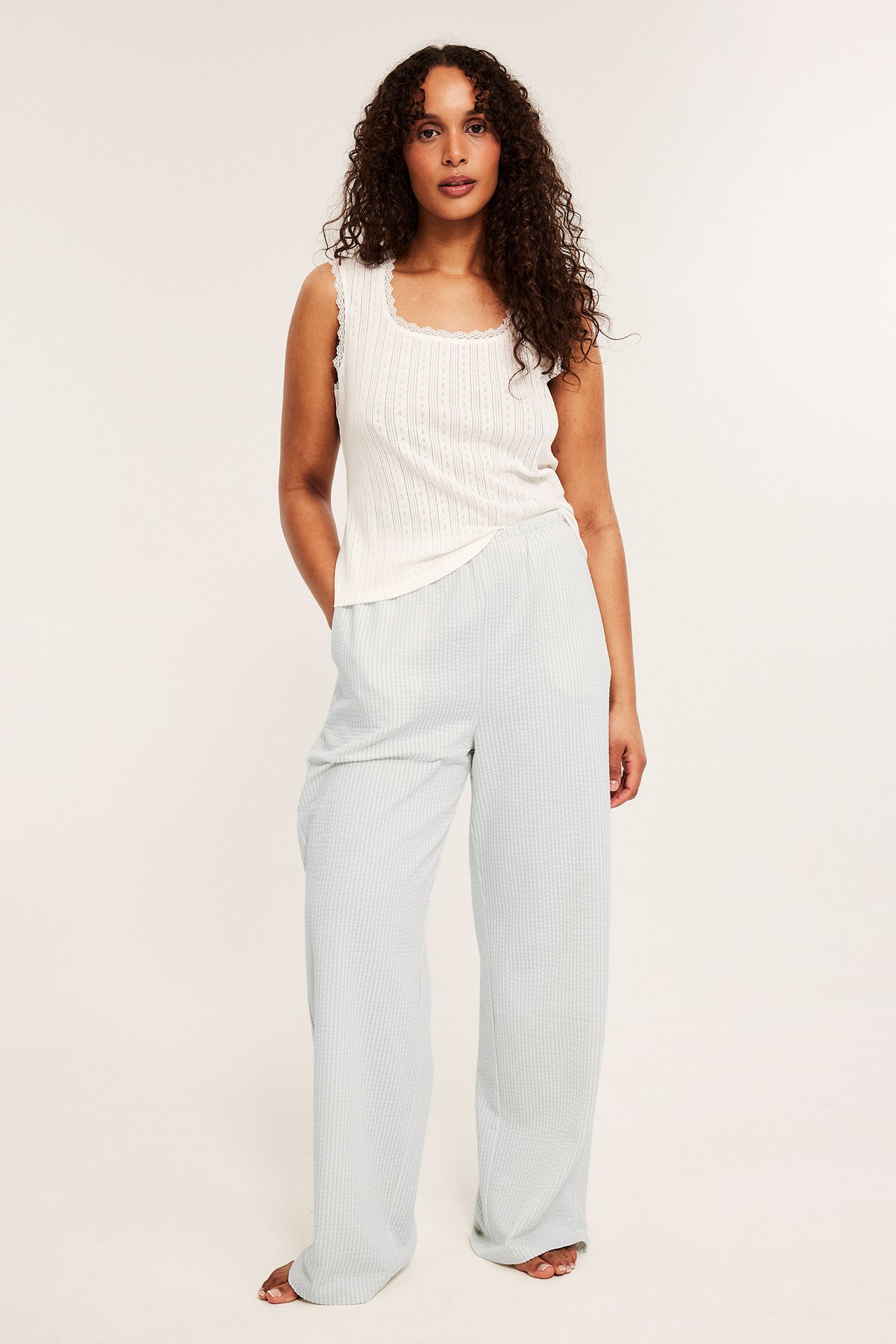 Framifrån: kvinna i ljusgröna randiga pyjamasbyxor med bäckebölja-struktur och vitt ärmlöst linne med spets.