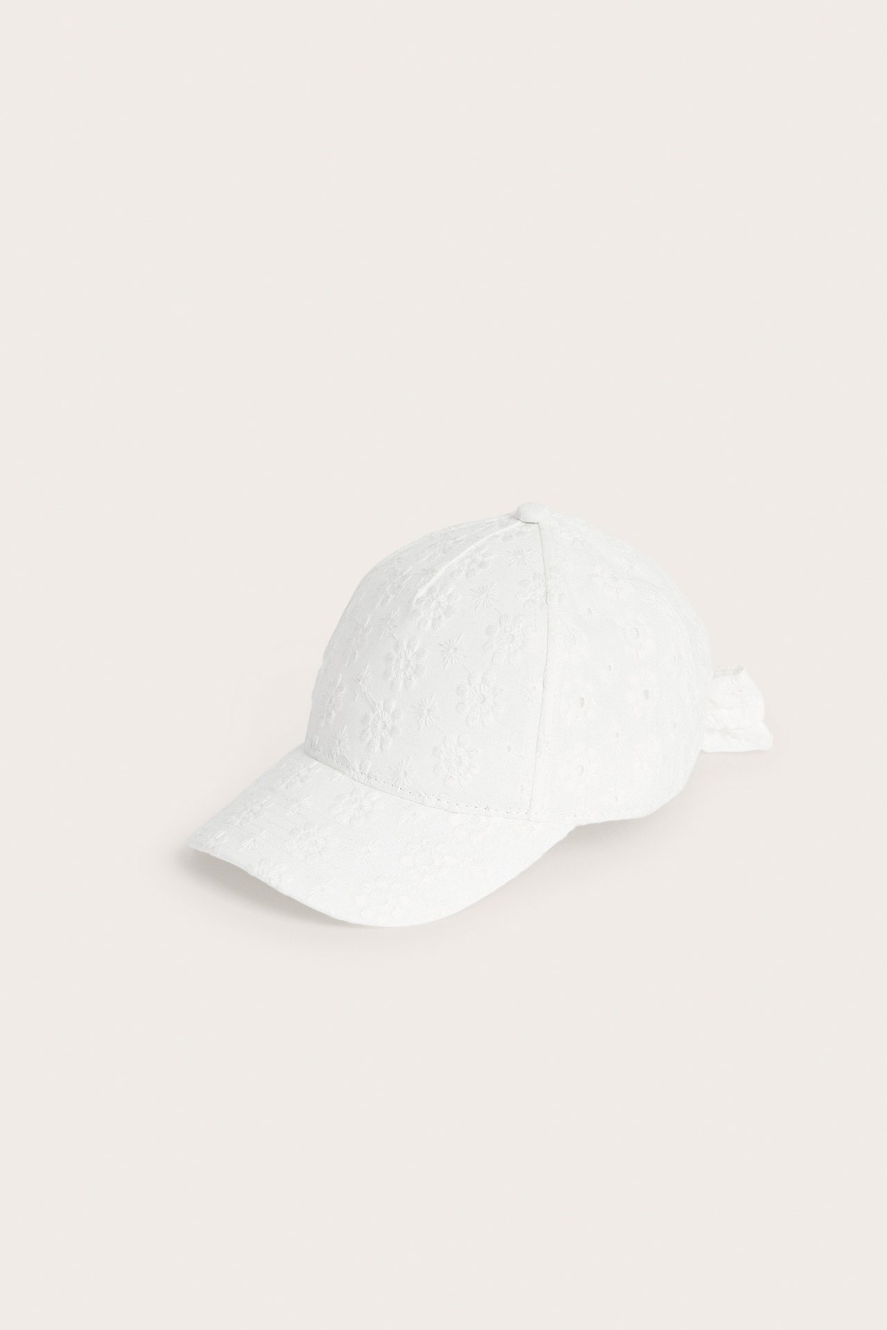 Offwhite broderie anglaise keps med strukturmönster och rosett baktill, front-sidovy.