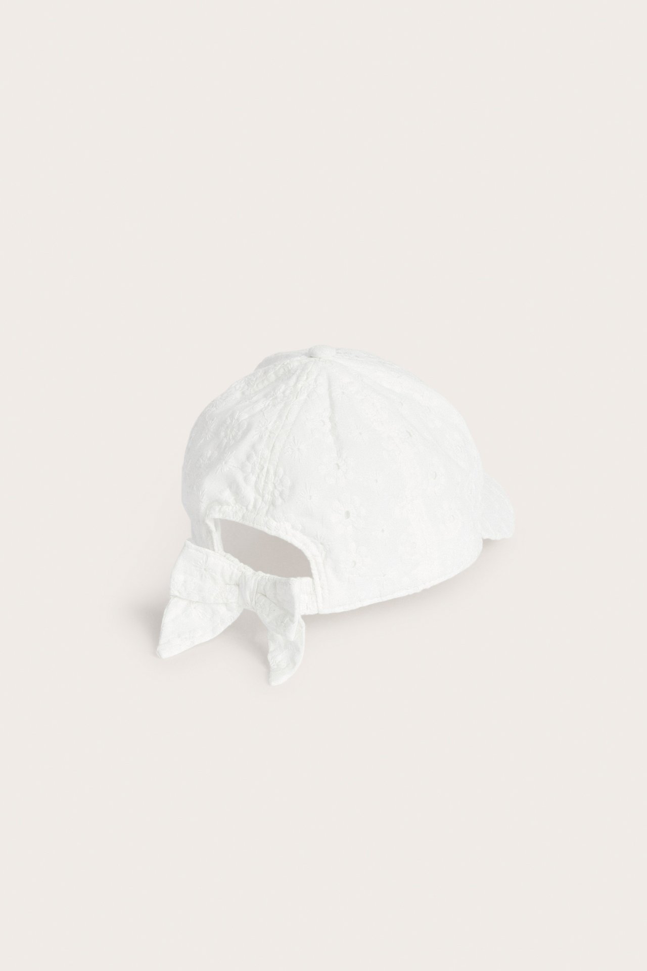 Bakre sidovy av en offwhite barnkeps med broderie anglaise-struktur och en rosett baktill.