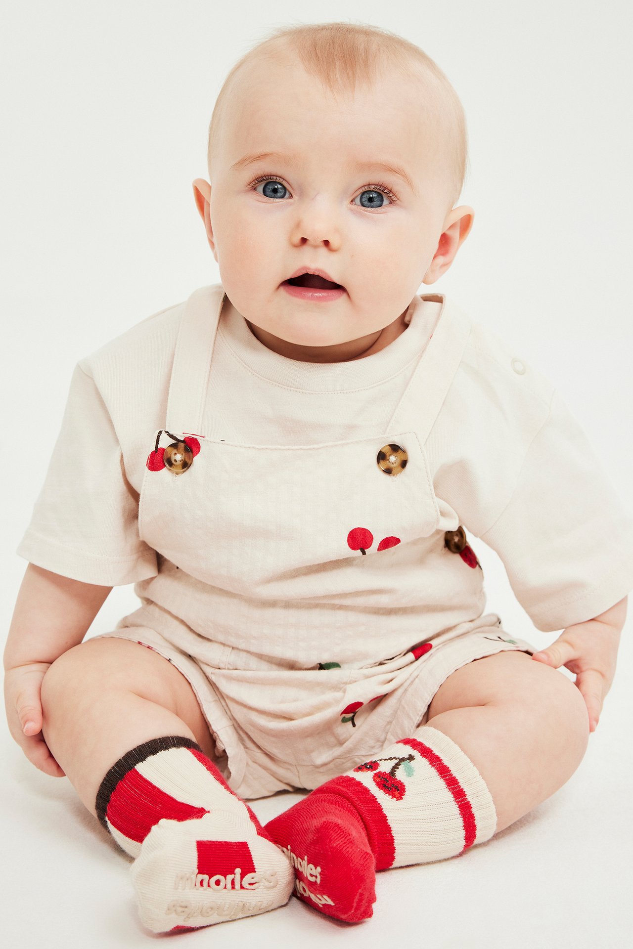 Frontvisning av baby i beige bomulls-shortsselebukse med kirsebærmønster, beige topp, røde/hvite sokker.