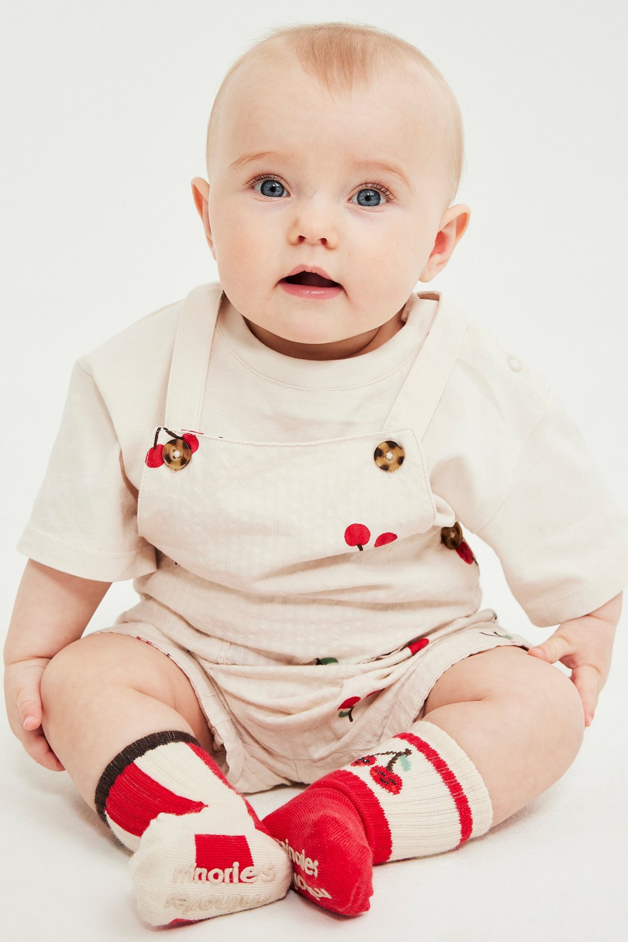 Widok z przodu dziecka w beżowych bawełnianych krótkich ogrodniczkach w wiśnie, beżowym topie, czerwono-białych skarpetkach.