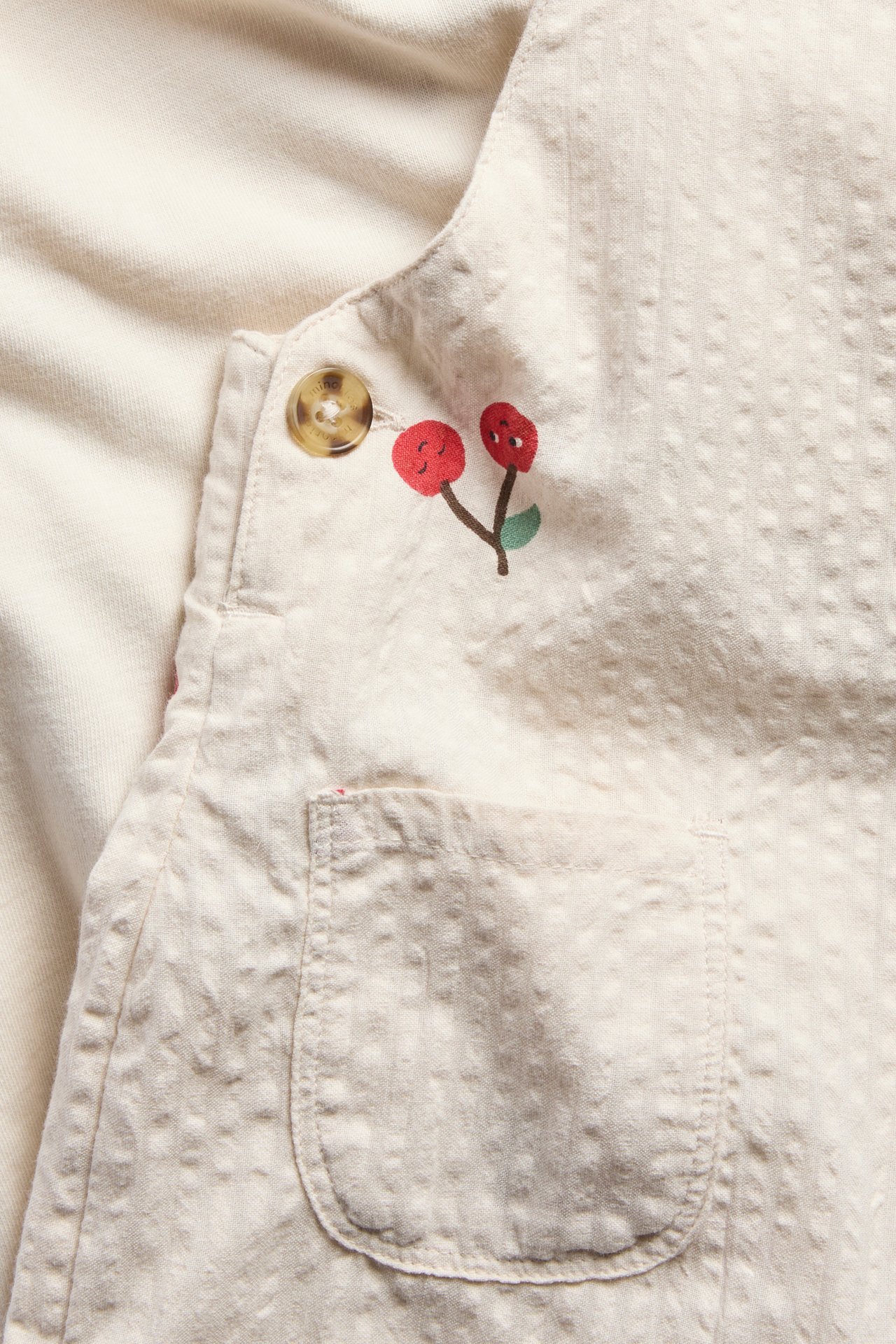 Lähikuva vauvan beigestä puuvillaisesta kuviollisesta haalarista, jossa kirsikkapainatus ja nappikiinnitys, yksivärisen paidan päällä.