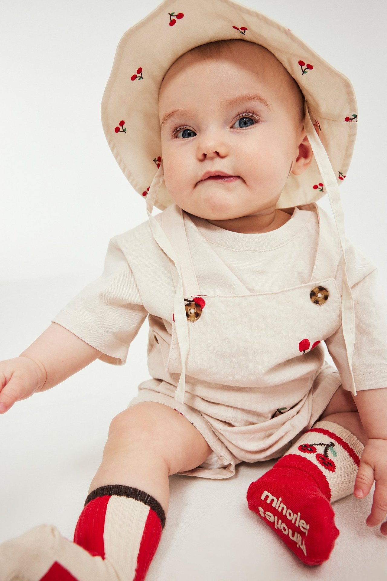 Frontvisning av baby i beige bomullssett med kirsebærmønster: bøttehatt, selebukse, stripete sokker.