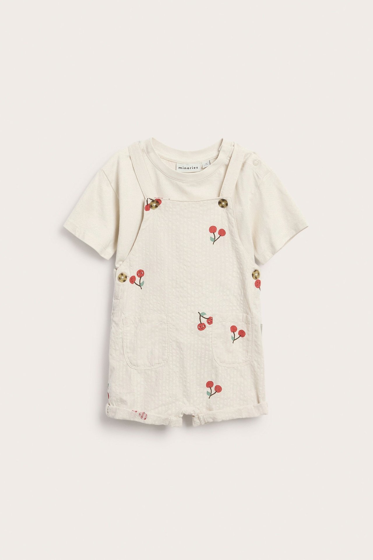 Framsida: Ett babyset med beige bomullsbyxor med rött körsbärsmönster, burna över en beige t-shirt.