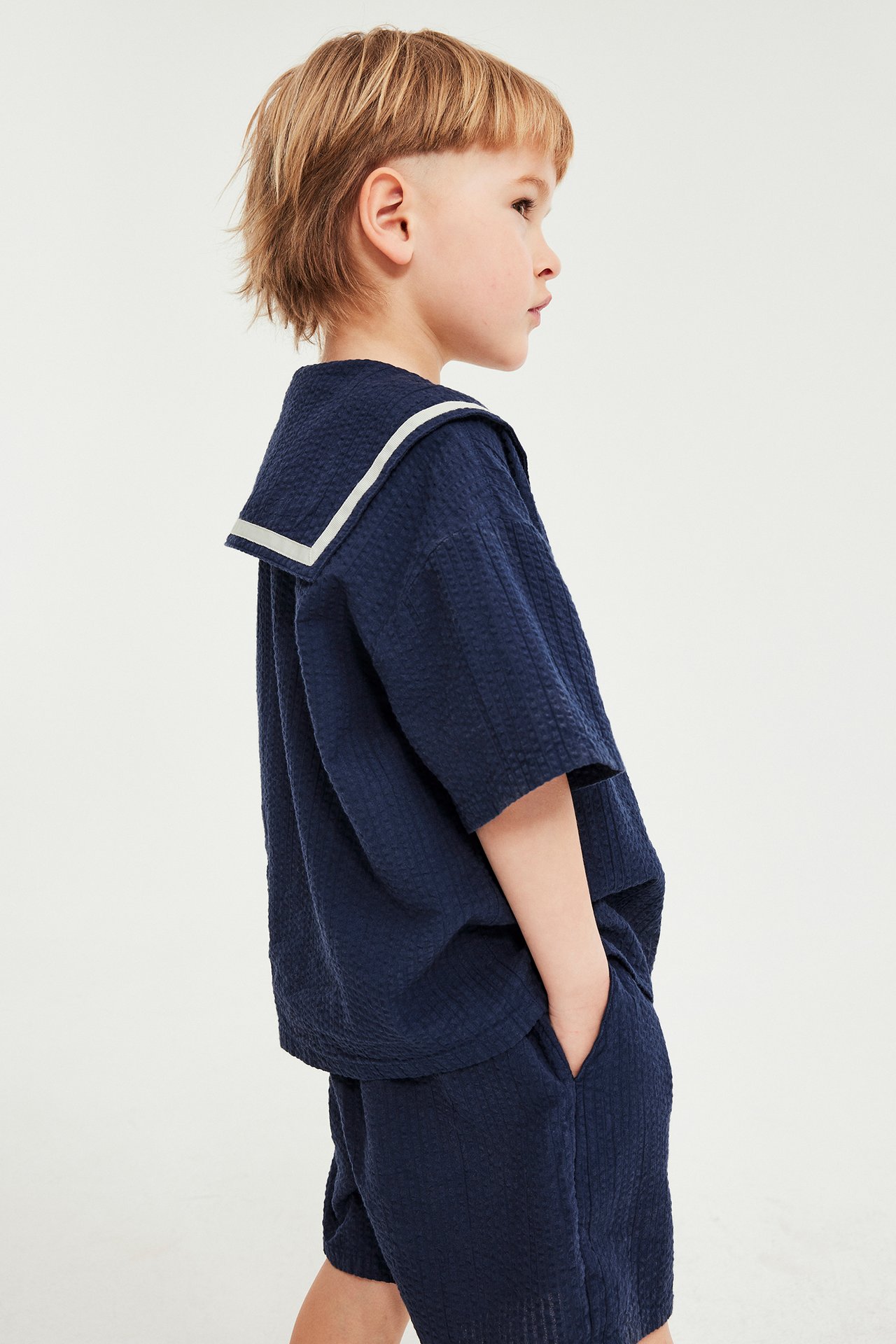 Sidebilde av barn i blå seersuckerskjorte med hvitstripet sjømannskrage og matchende blå shorts.