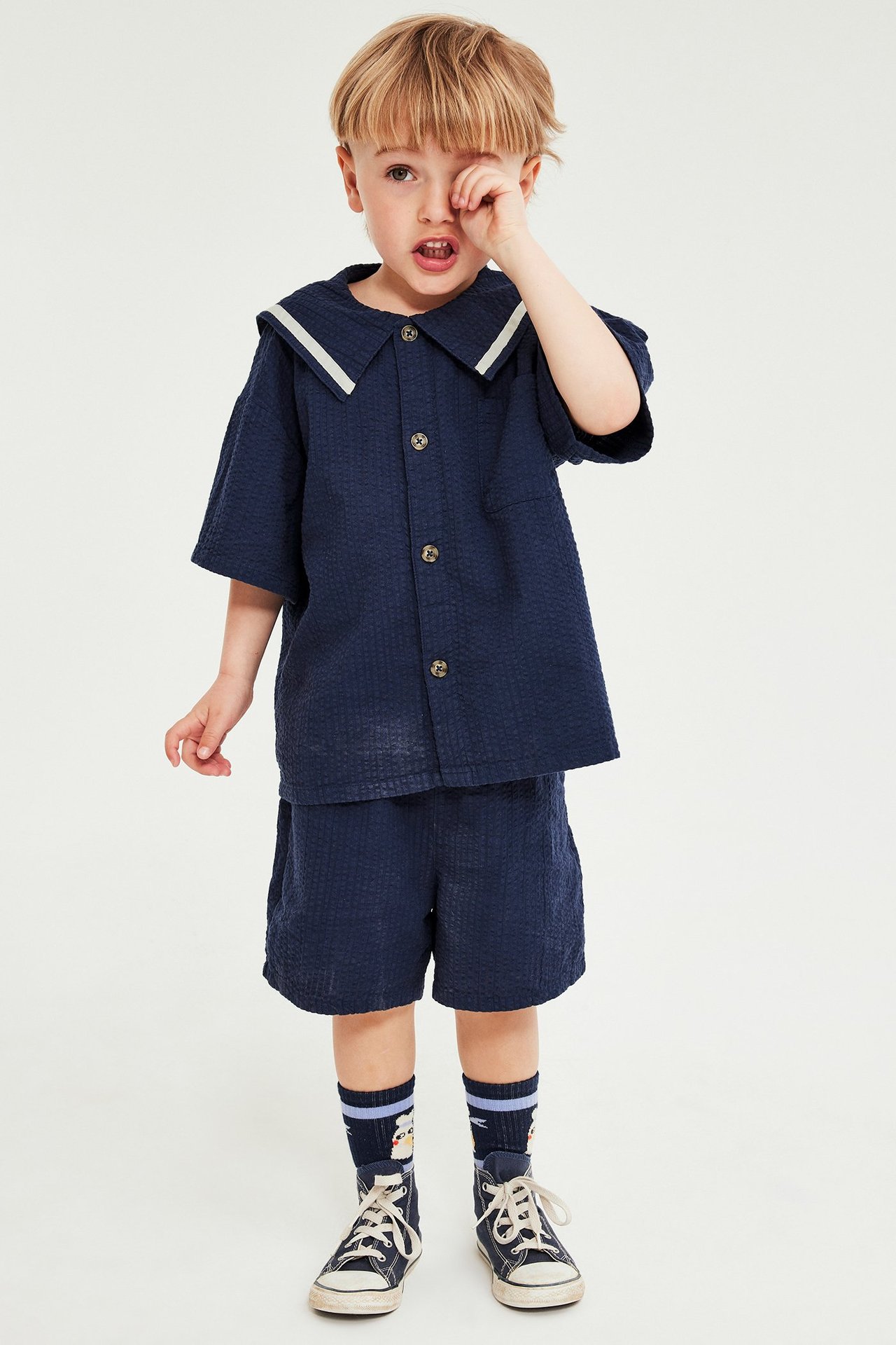 Framifrån, ett barn i blå seersucker skjorta med sjömanskrage, matchande shorts, mörkblå strumpor och sneakers.