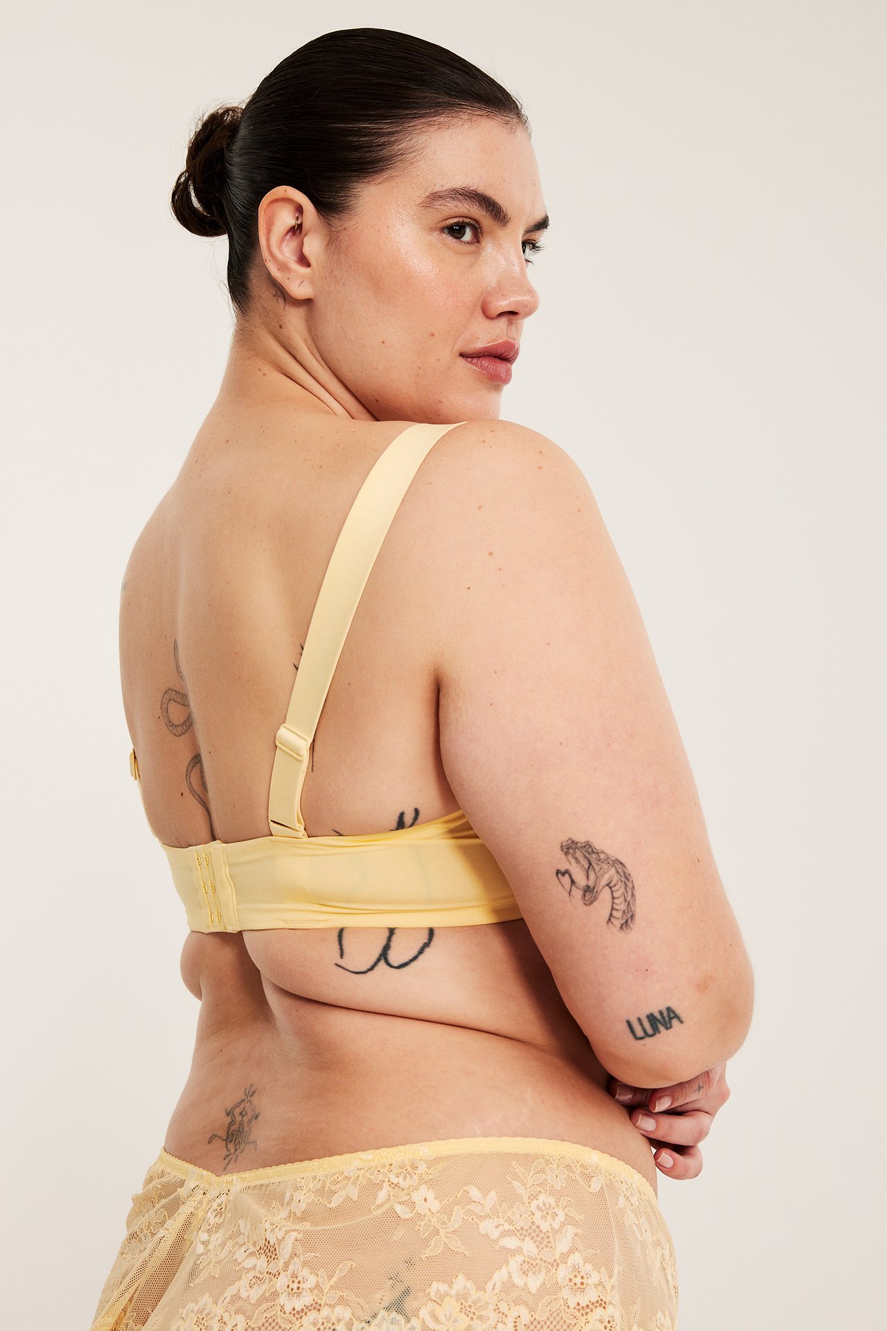 Naisen takasivuprofiili yllään vaaleankeltainen pitsikaariliivi ja yhteensopivat pitsialushousut.