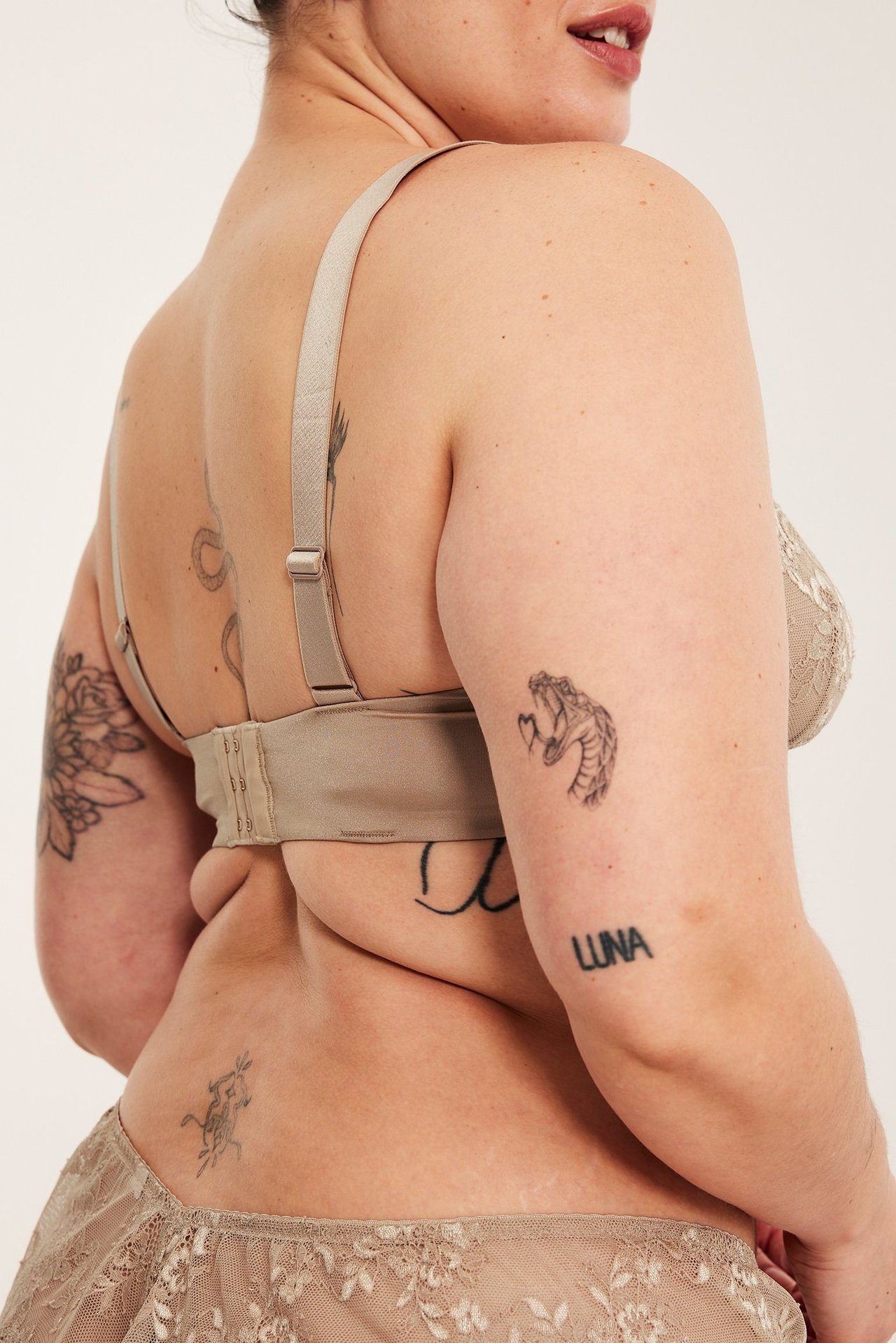 Nainen takaapäin yllään beige pitsinen kaarituellinen rintaliivi ja yhteensopivat pikkuhousut.