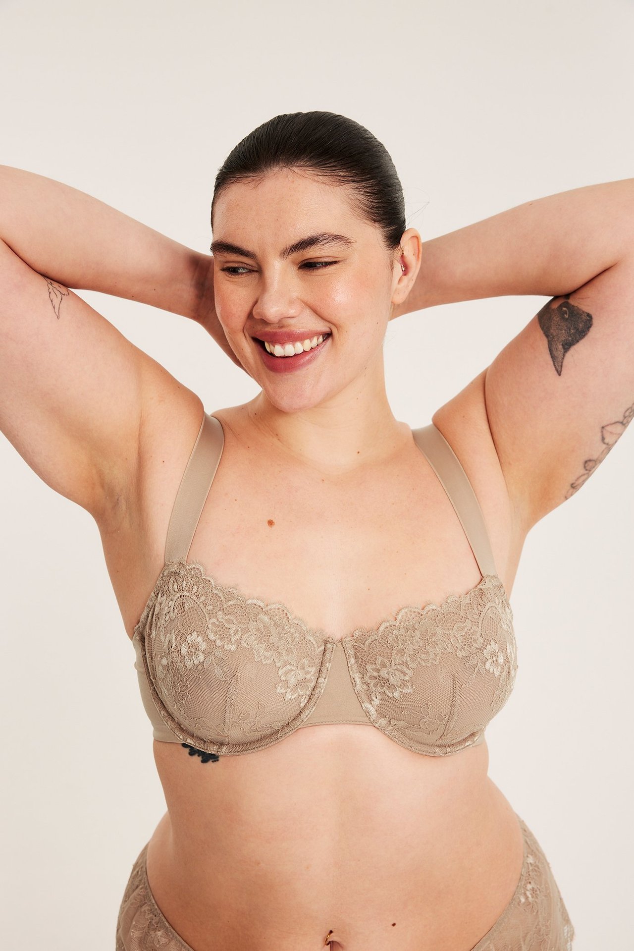 Framifrån, leende kvinna i ljusbrun spets-bh med bygel och matchande spetstrosa.