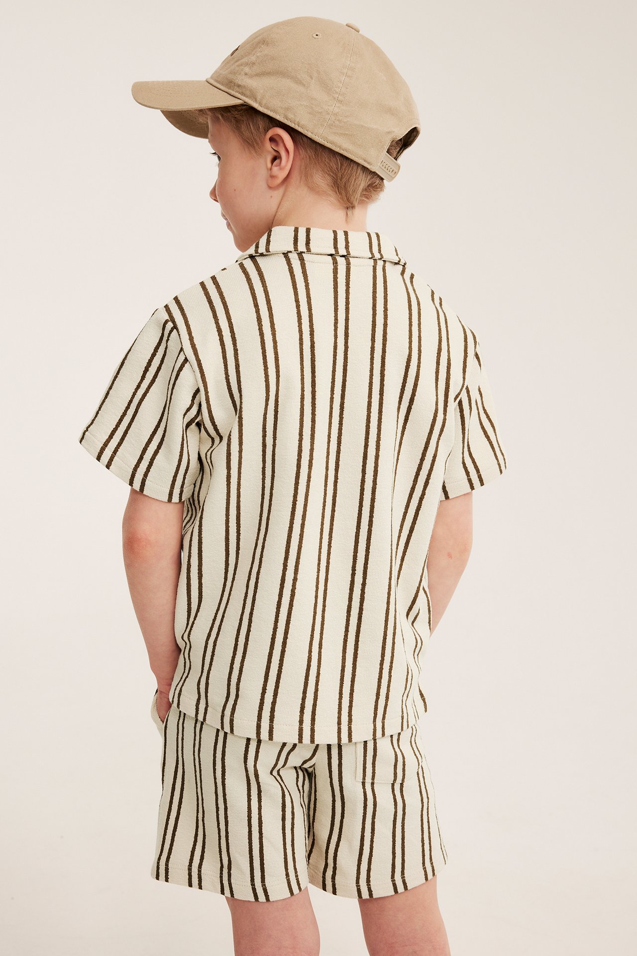 Bakfra av et barn i lys beige og brunstripete jerseyshorts, matchende skjorte og beige caps.