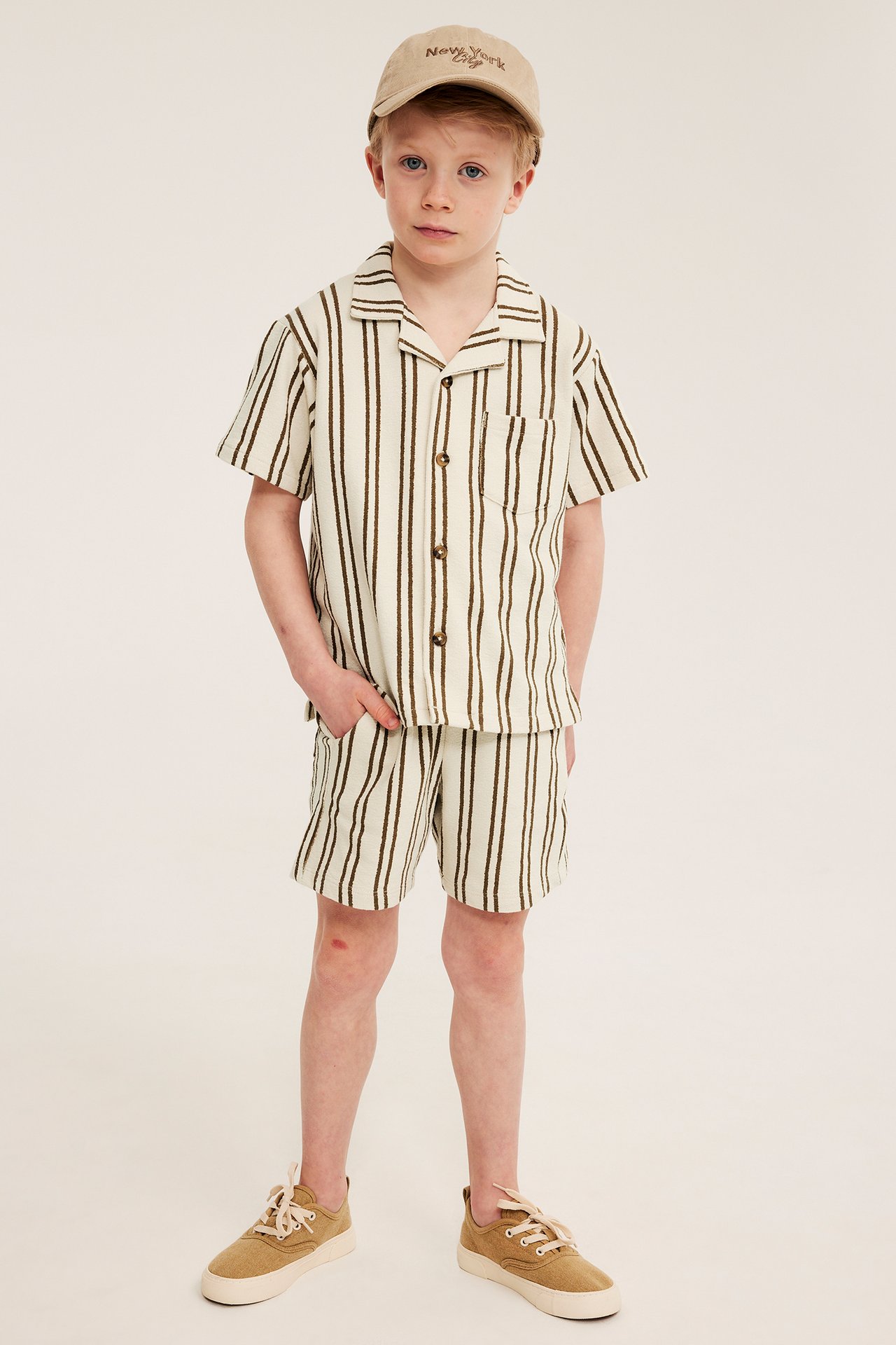 Frontutsikt: Barn i lysebeige stripete bomullsjersey-shorts og matchende skjorte, med beige caps og joggesko.