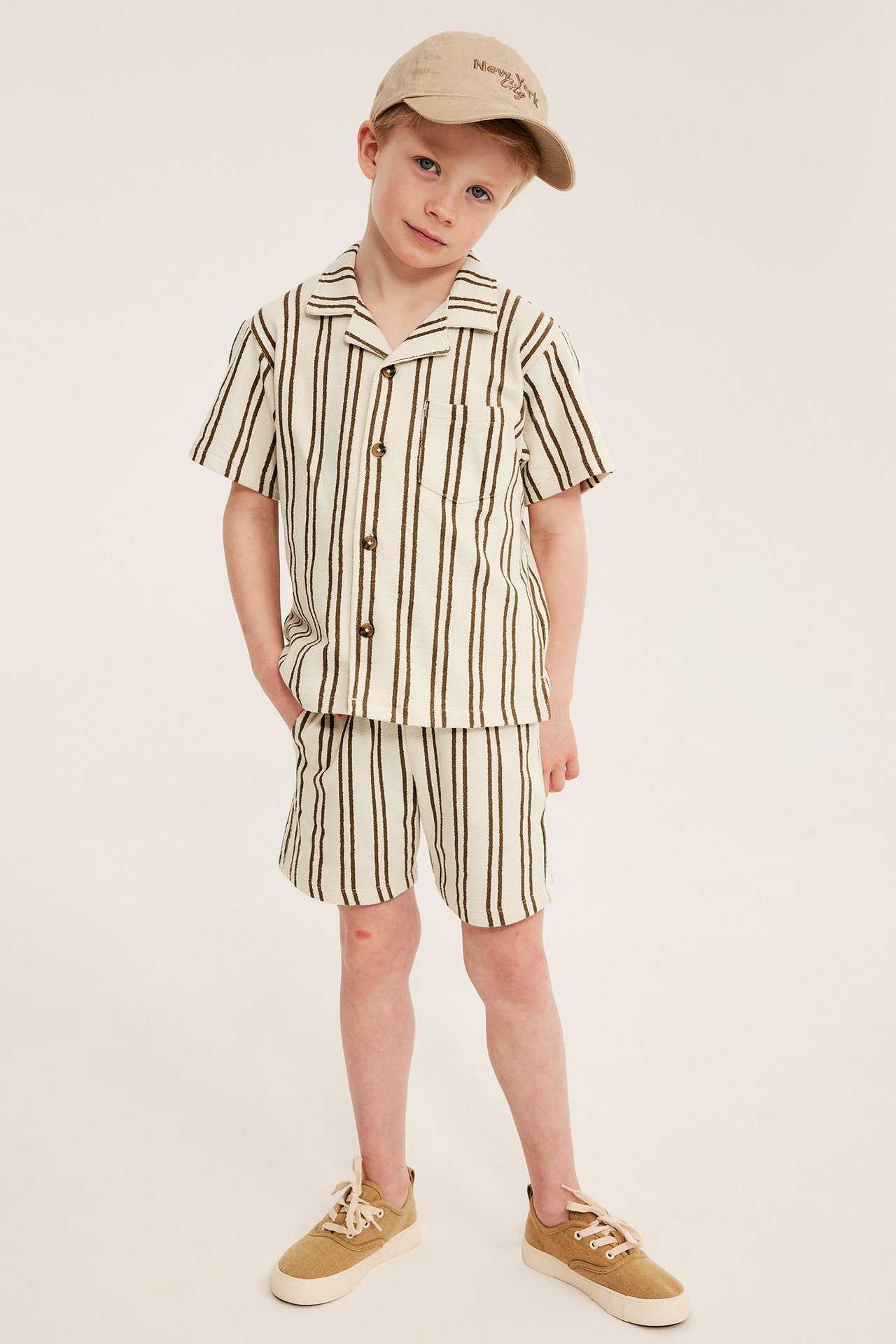 Lapsi: vaaleanbeiget raidalliset shortsit, yhteensopiva paita, beige lippis ja tennarit.