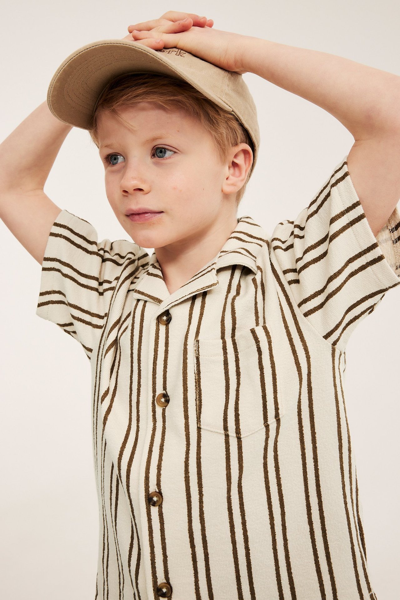 Dziecko w jasnobeżowej koszuli w paski z krótkim rękawem, guzikami i beżowej czapce z daszkiem.