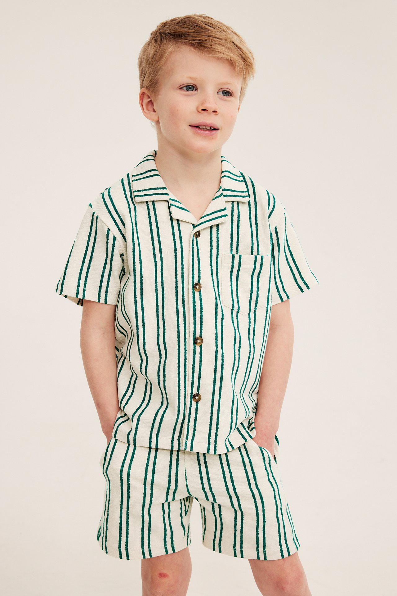 Forfra, en gutt i grønn- og kremhvite stripete jersey-shorts og matchende kortermet skjorte.