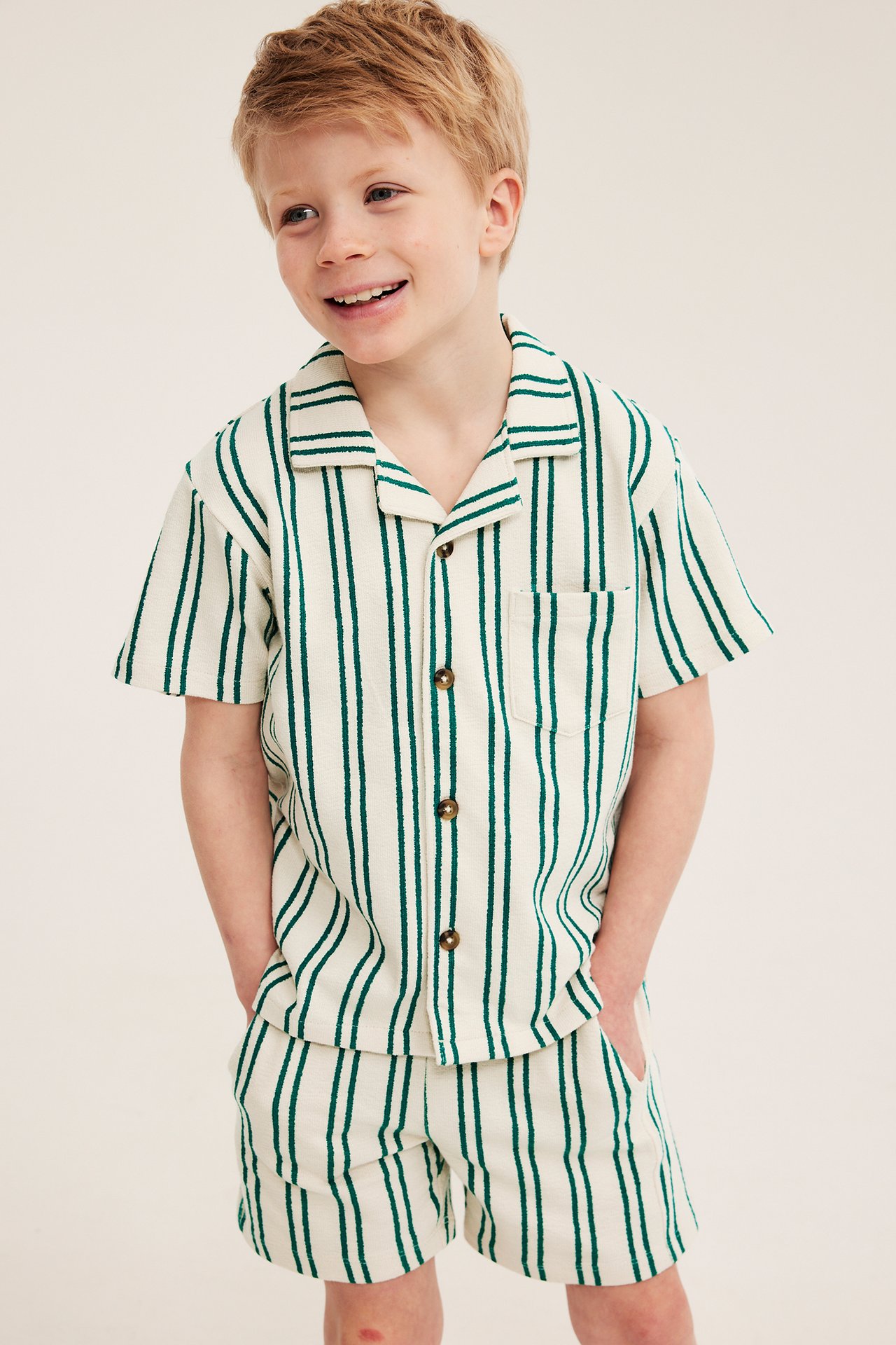 Widok z przodu uśmiechniętego chłopca w kremowo-zielonej koszulce frotte w paski i pasujących szortach.