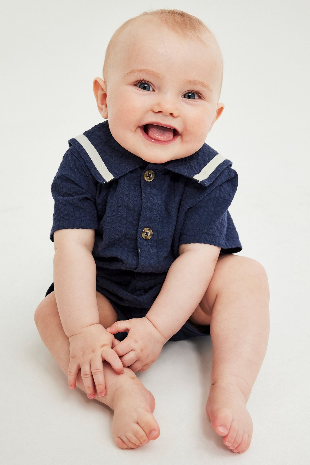 Frontvy av en glad baby i marinblå seersucker kortärmad romper med randig sjömanskrage och knappar fram.