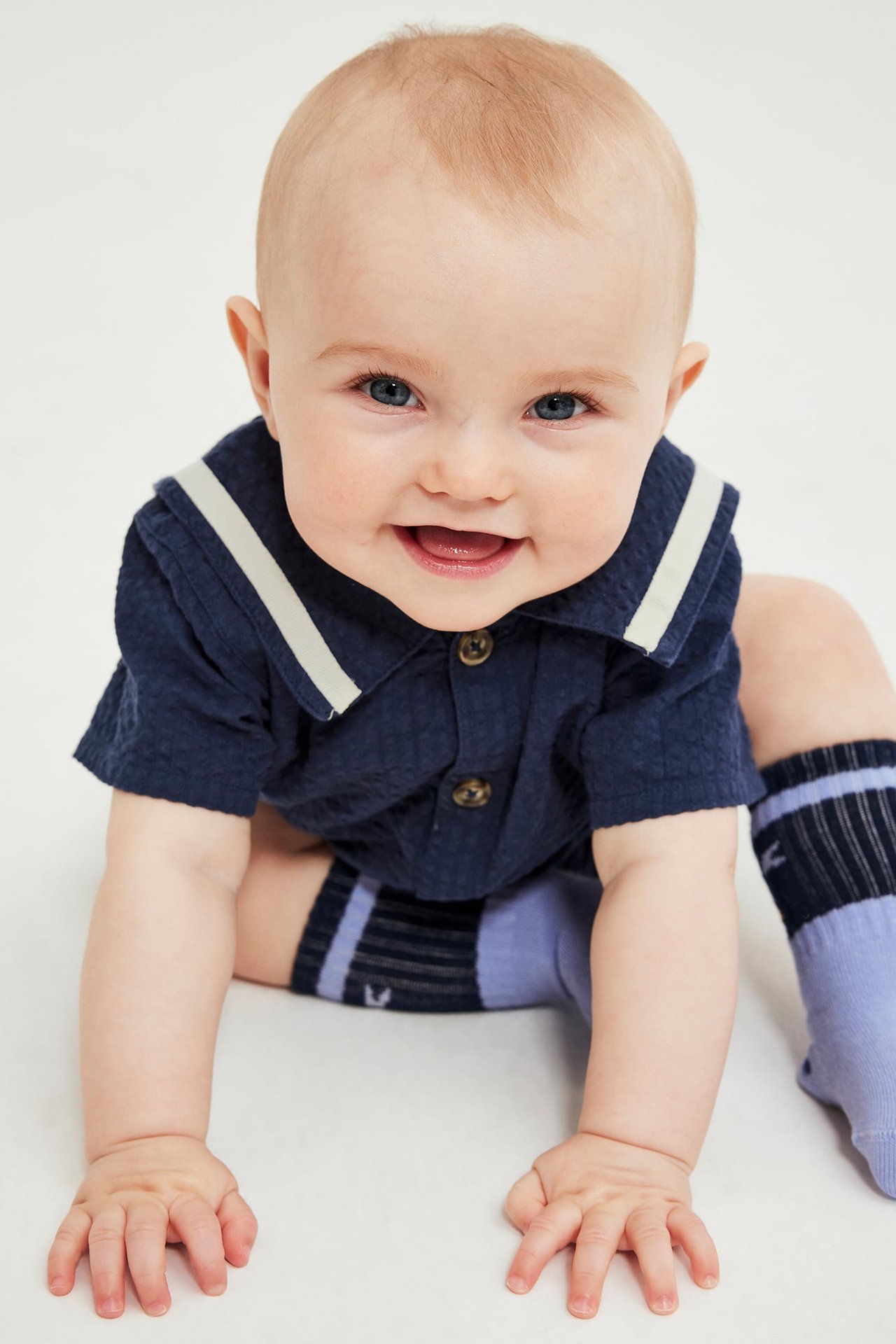 Frontvy av en leende baby i blå kortärmad romper med randig krage och ljusblå randiga strumpor.