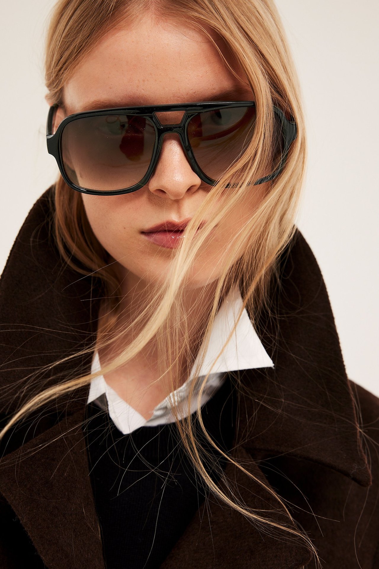 Lähikuva edestä naisesta, jolla mustat pilotsolmilasit, valkoinen paita ja ruskea takki.