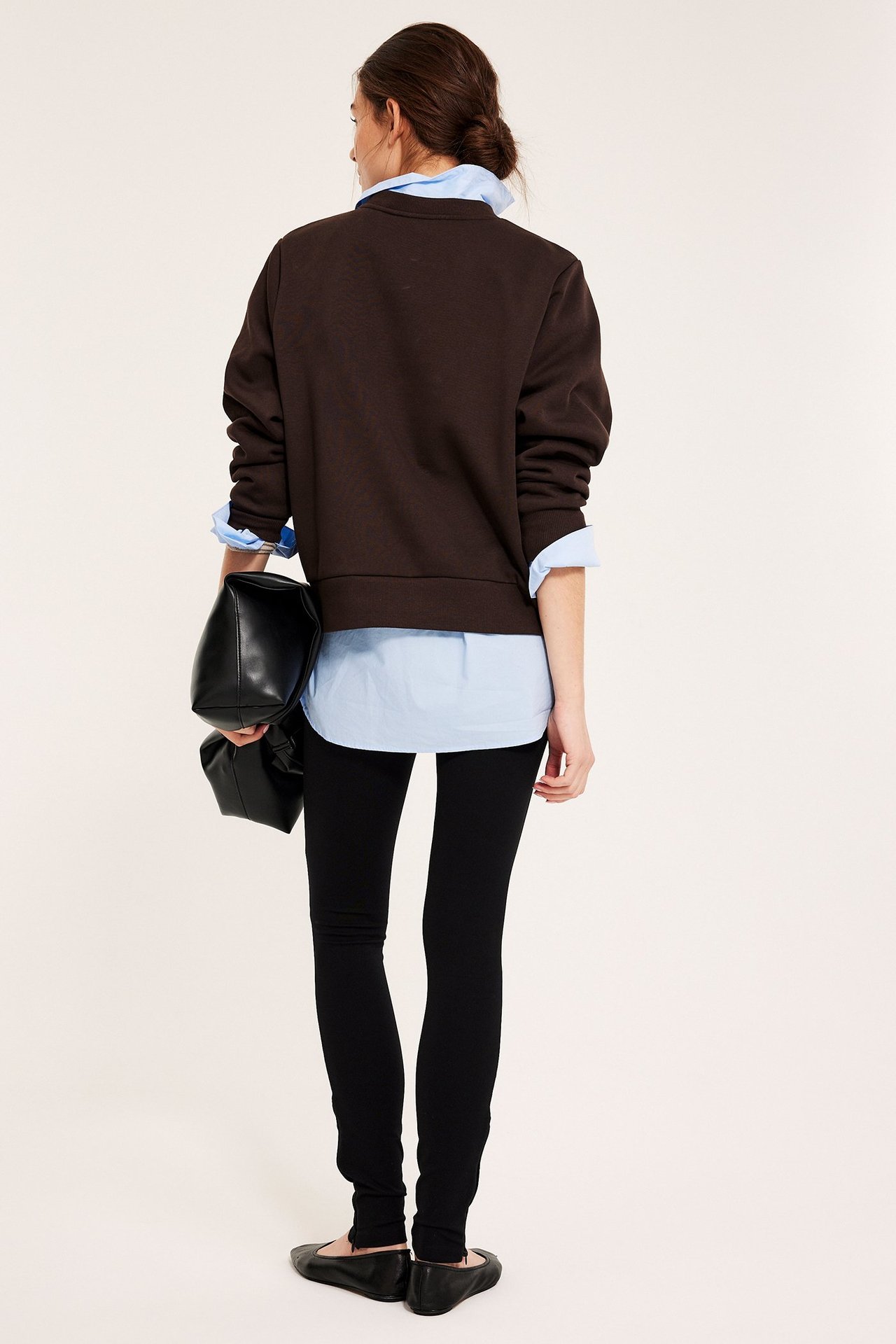 Takakuva naisesta tummanruskeassa collegepaidassa vaaleansinisen paidan päällä, mustissa leggingseissä ja kengissä, pitäen mustaa käsilaukkua.