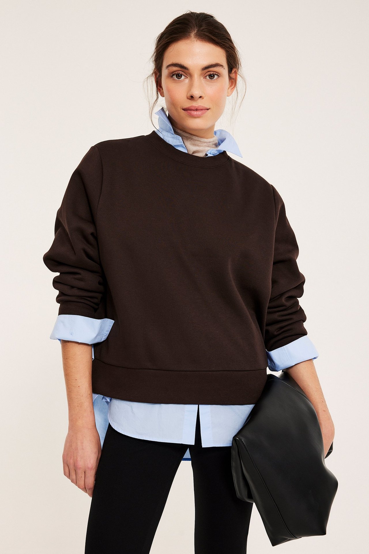 Edestä: Nainen tummanruskea collegepaita, vaaleansininen paita, mustat housut, pitää mustaa clutchia.