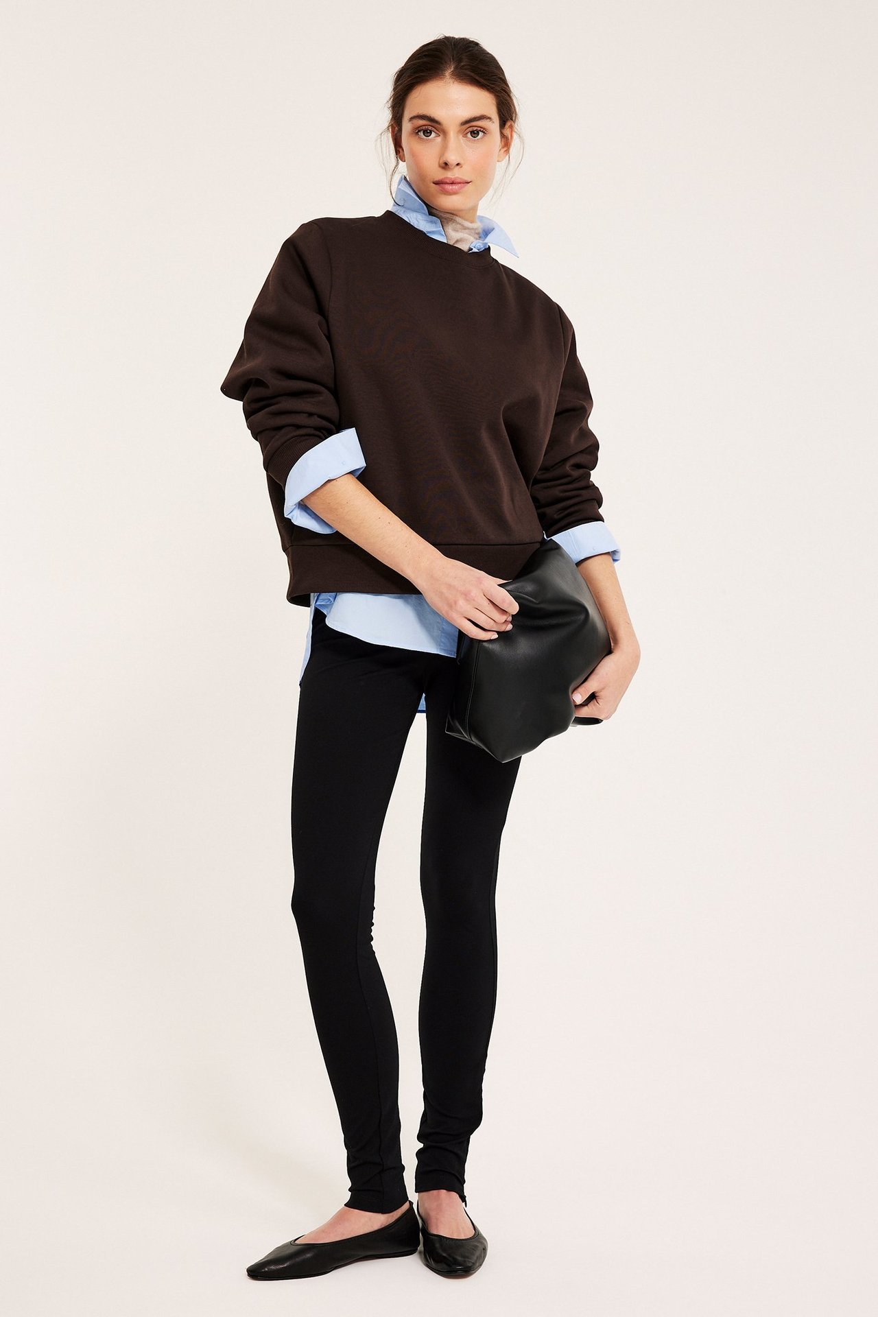 Edestä: Nainen tummanruskeassa collegepaidassa, vaaleansininen paita alla, mustat leggingsit, kengät, musta käsilaukku.