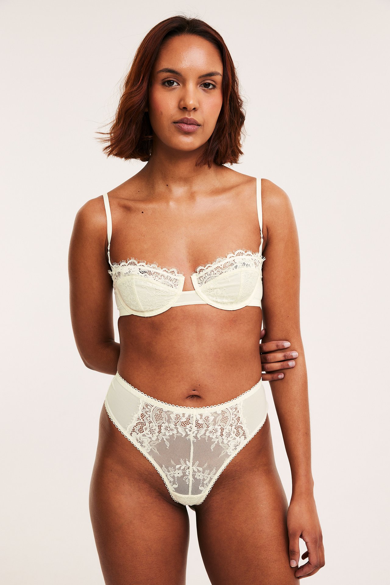 Frontvy av en kvinna i en offwhite spetsstringtrosa och matchande spets-bh.