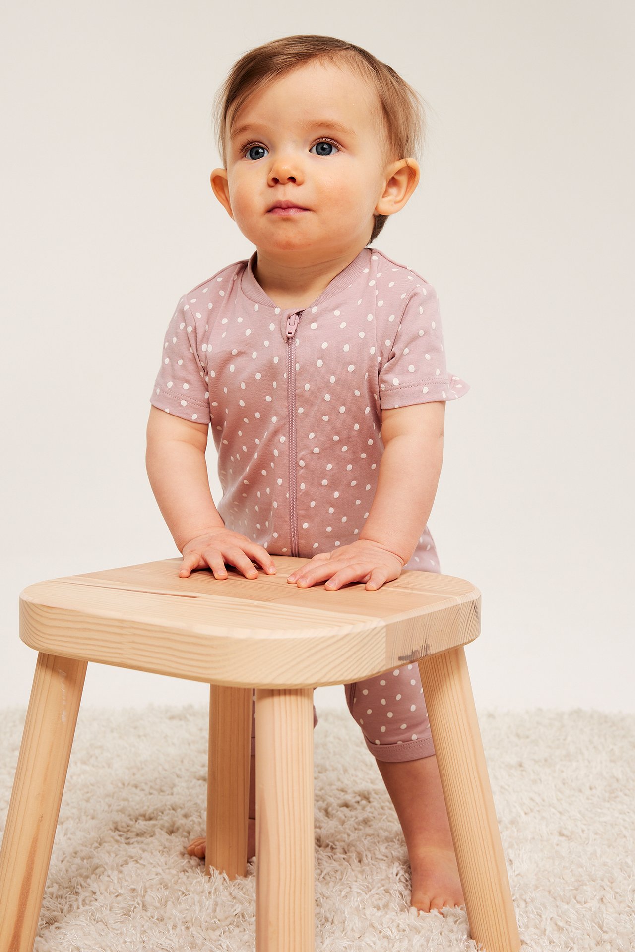 Dziecko w pudrowo-różowym pajacyku z krótkim rękawem, kropkami ecru i zamkiem, trzyma drewniany taboret.
