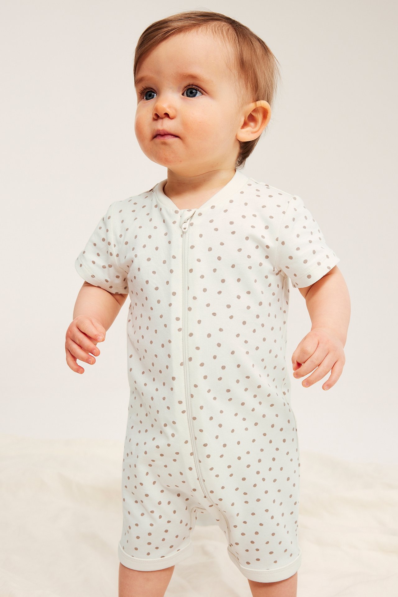Widok z przodu: niemowlę w beżowym kombinezonie z krótkim rękawem, w brązowe kropki, z zamkiem błyskawicznym.