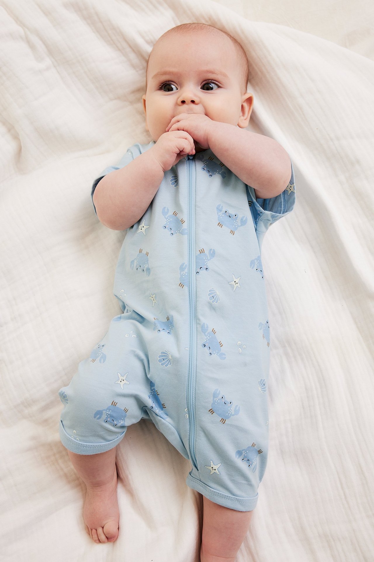 Ylhäältä kuvattuna, vauva yllään vaaleansininen lyhythihainen pyjama, jossa on rapukuvio ja etuvetoketju.