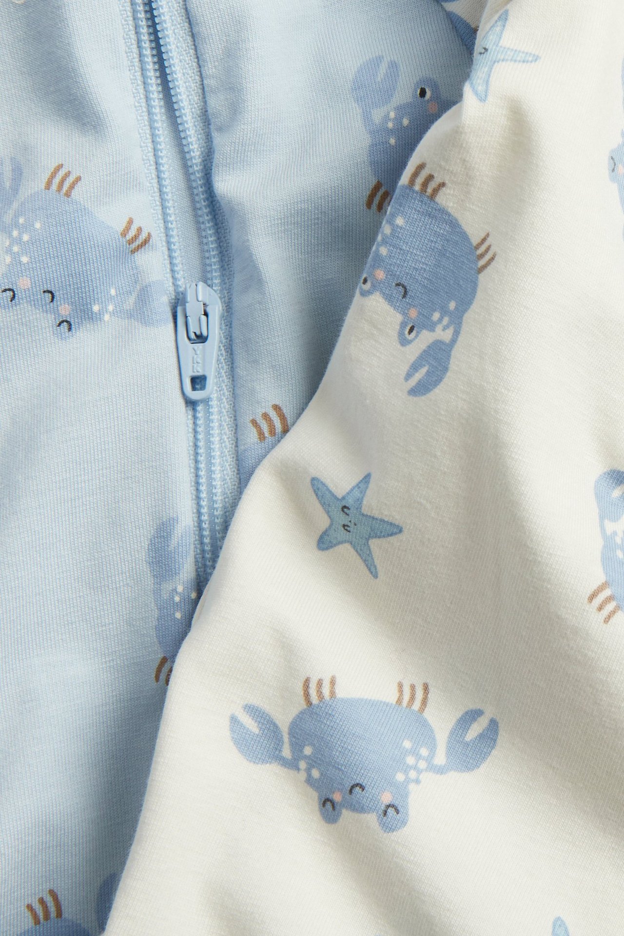 Zbliżenie na jasnoniebieski pajacyk niemowlęcy z zamkiem i nadrukiem krabów, obok białego pajacyka.