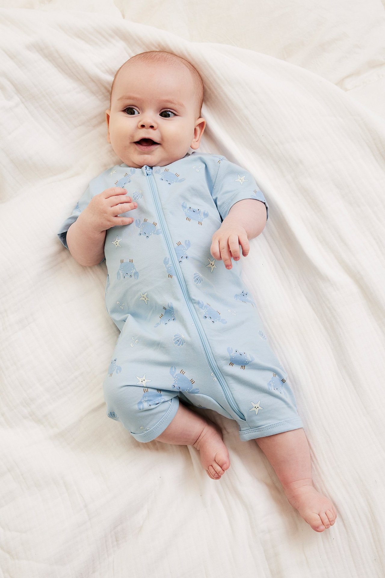 Niemowlę w jasnoniebieskim krótkim pajacyku w kraby z zamkiem z przodu, widok z góry.
