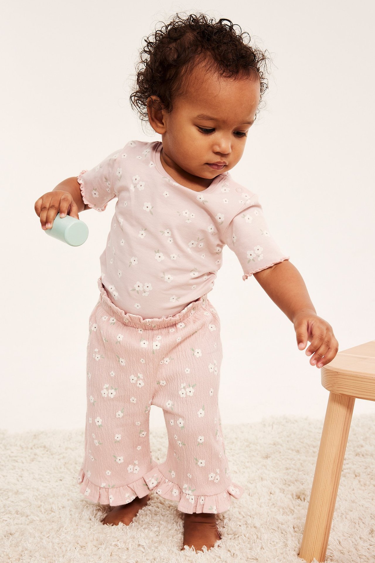 Niemowlę w różowym body z krótkim rękawem w kwiaty, pasujących spodniach z falbanami, trzyma zieloną zabawkę.