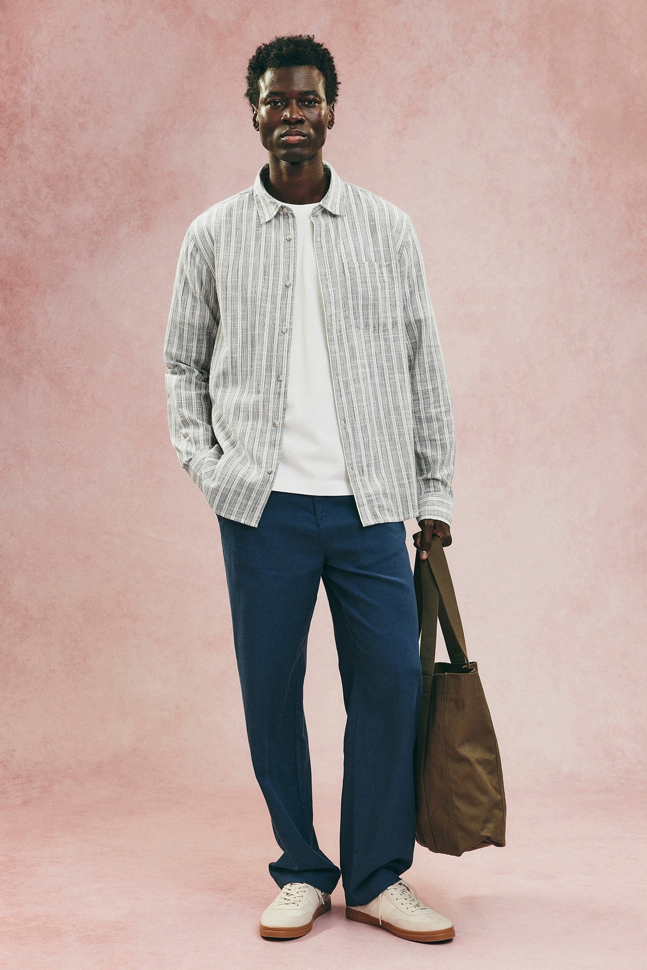 Mężczyzna w otwartej niebieskiej koszuli w paski, białym T-shircie, niebieskich spodniach, białych sneakersach, trzyma zieloną torbę.