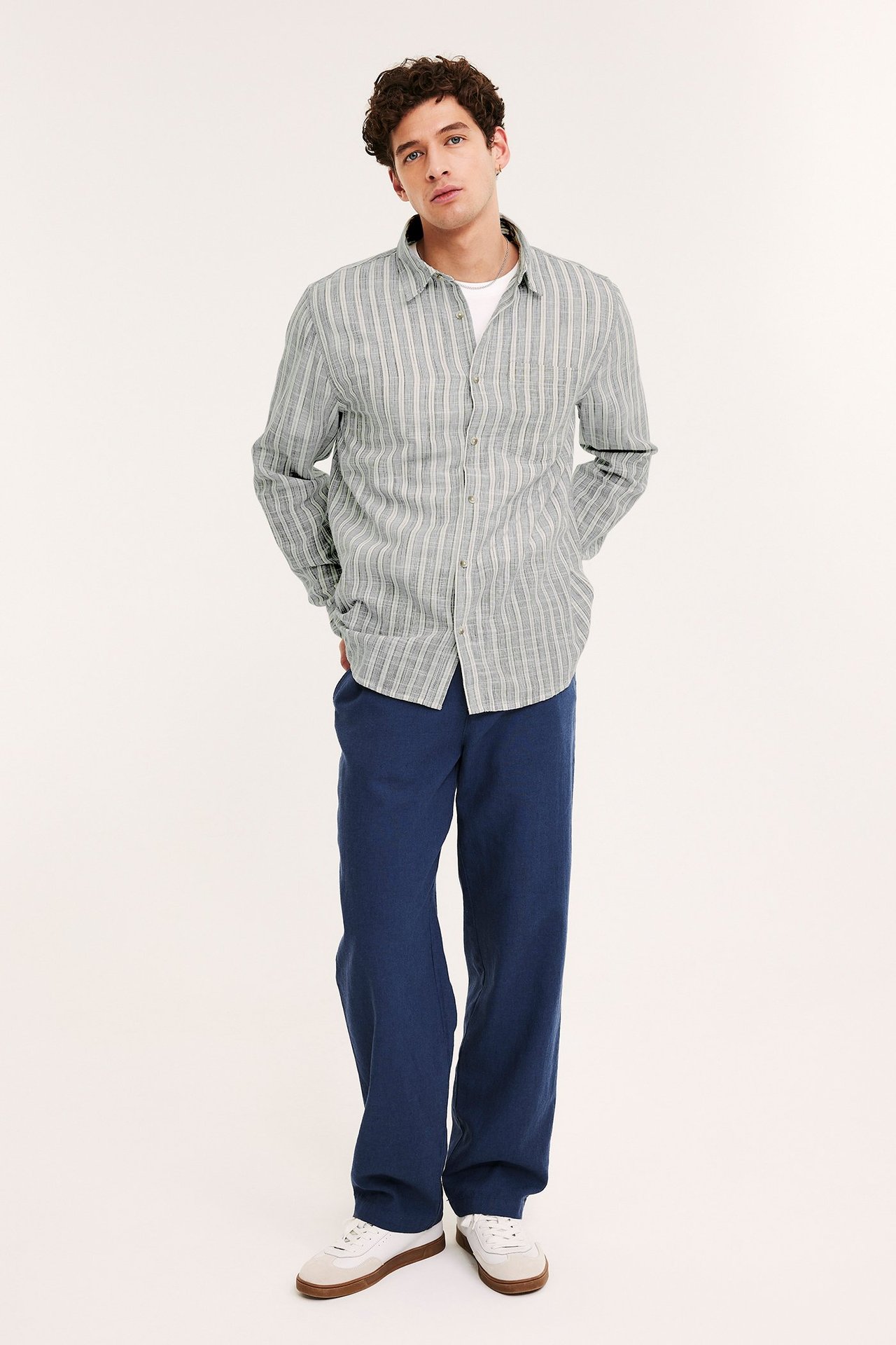 Edestäpäin mies yllään sinivalkoraidallinen puuvillapaita, valkoinen t-paita ja tummansiniset housut.