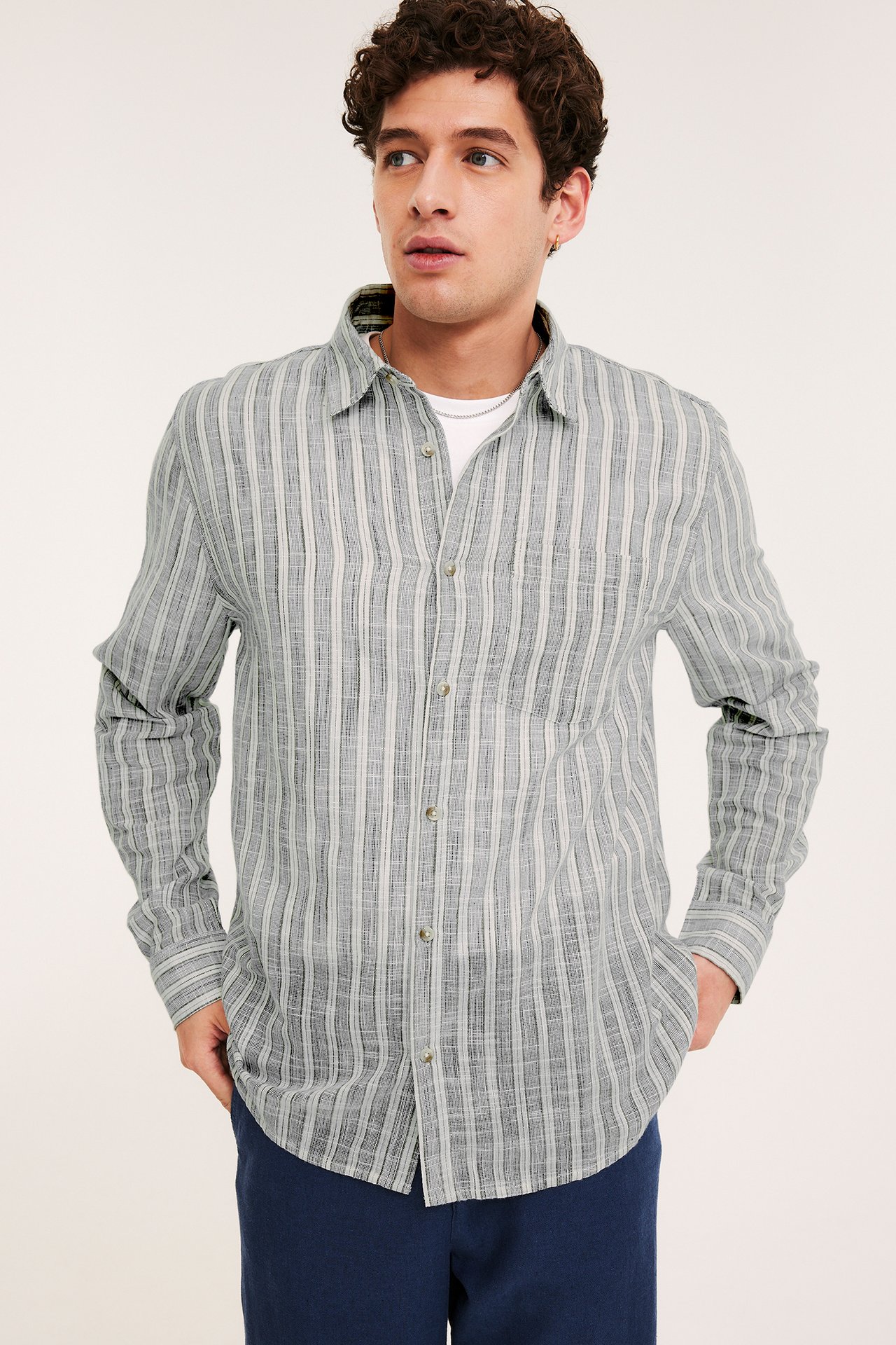 Mies edestäpäin, siniraidallinen puuvillapaita, avoin valkoisen t-paidan päällä ja tummansiniset housut.