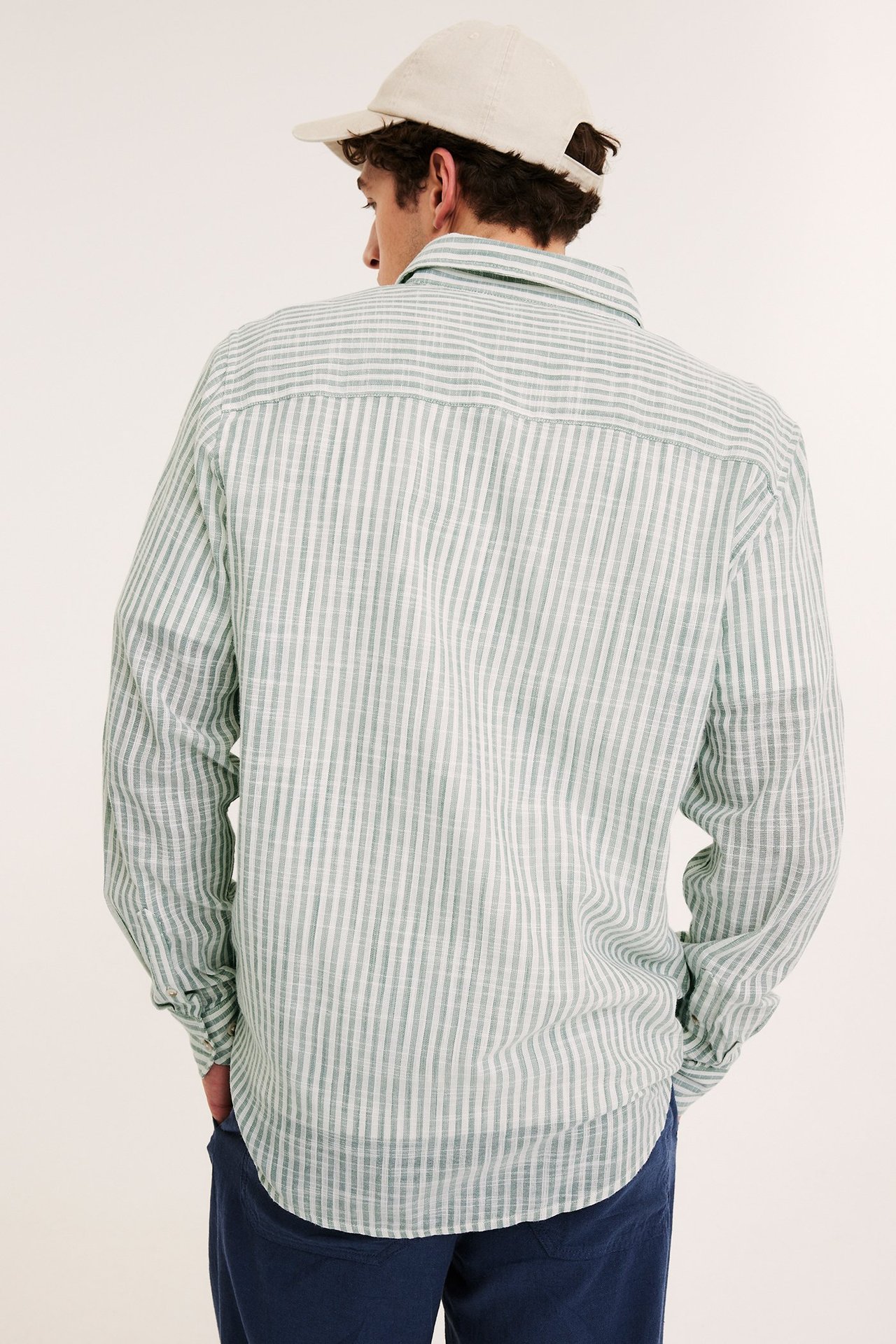 Bakfra av en mann i lys grønn- og hvitstripet bomullsskjorte, beige caps og mørkeblå bukser.